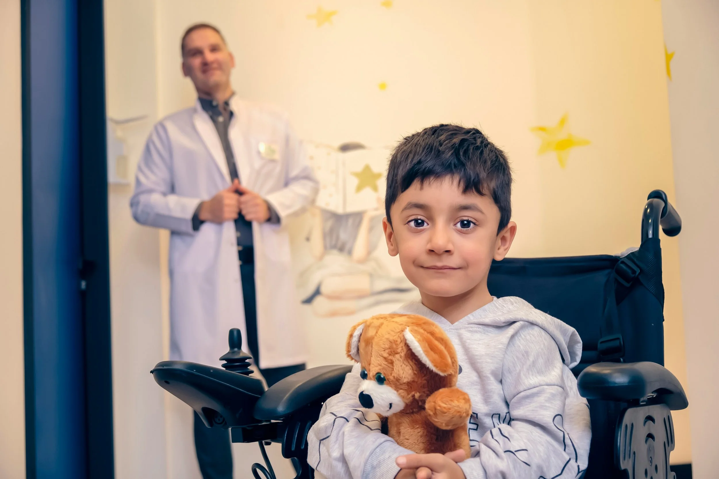 A young boy sitting in a wheelchair, holding a teddy bear, with a doctor standing in the background near a hospital wall decorated with stars.