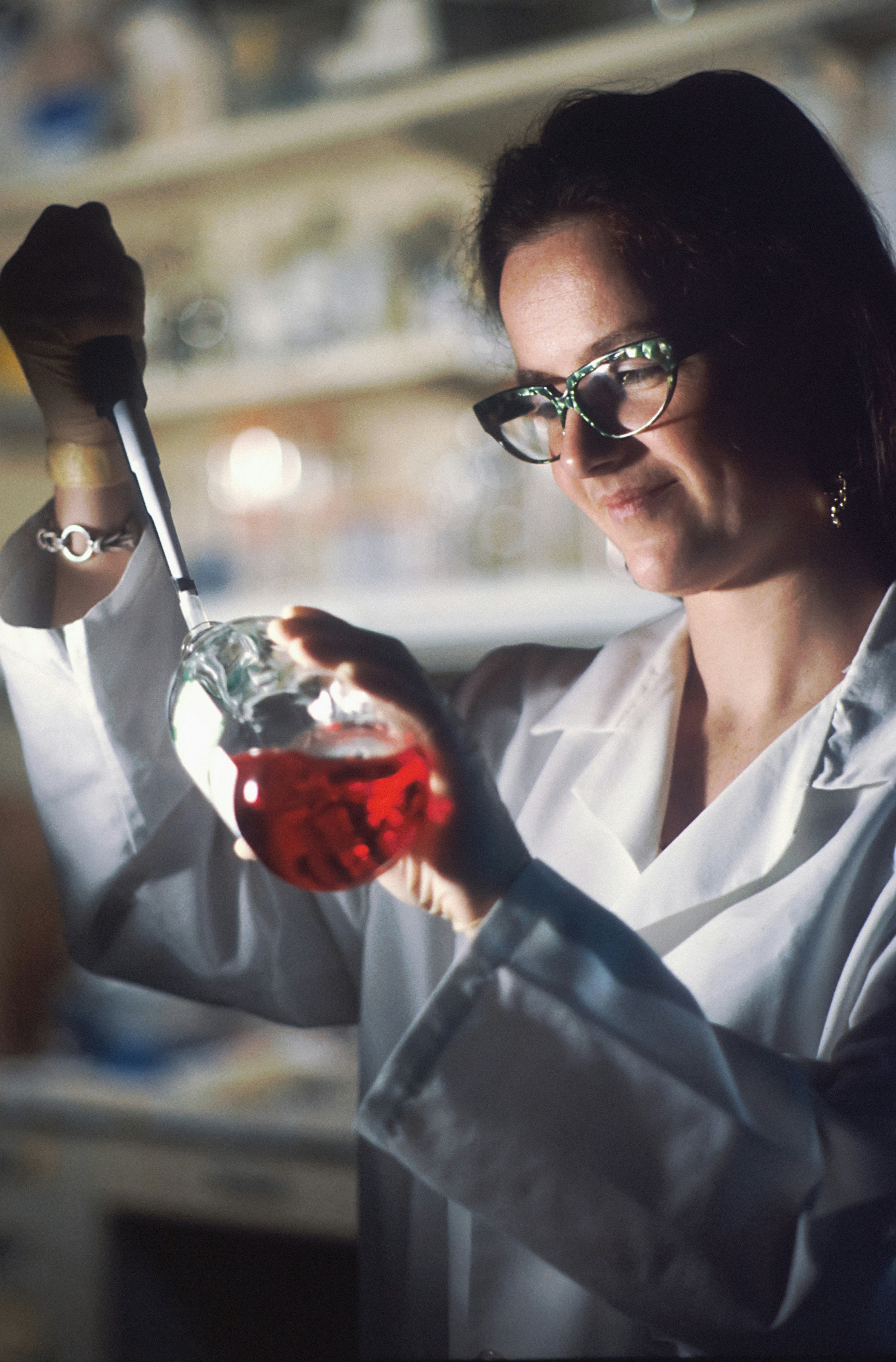 A woman with dark hair wearing glasses and a white lab coat holds a glass beaker filled with red liquid in one hand and a pipette in the other, working in a laboratory.