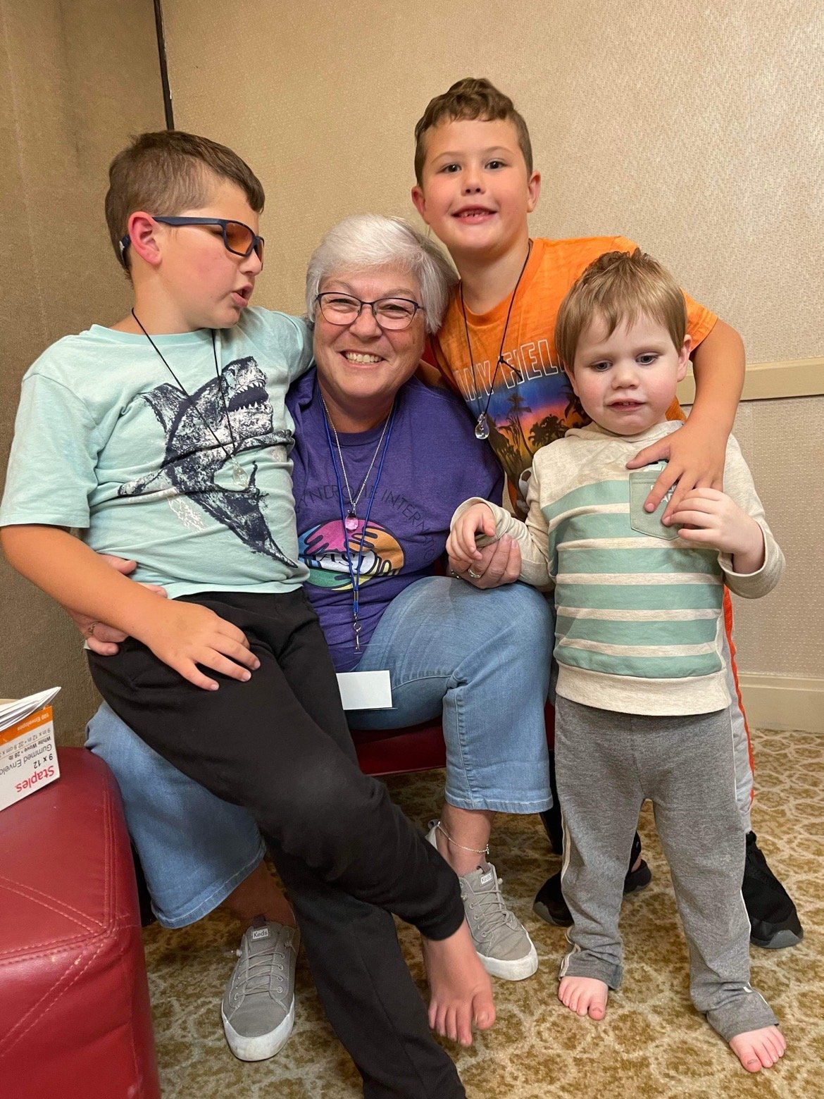 A smiling older woman with white hair and glasses sits in the center, surrounded by three young boys, in a room with beige walls and carpeted floor.