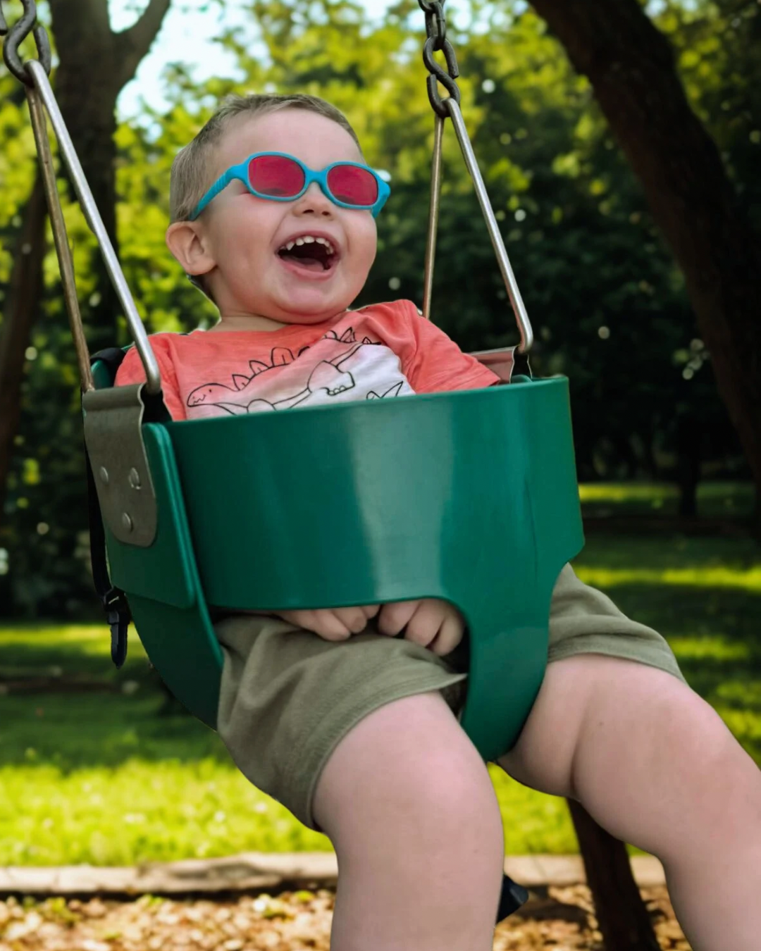A young child in sunglasses sitting in a green outdoor swing, smiling happily in a park with trees and grass.
