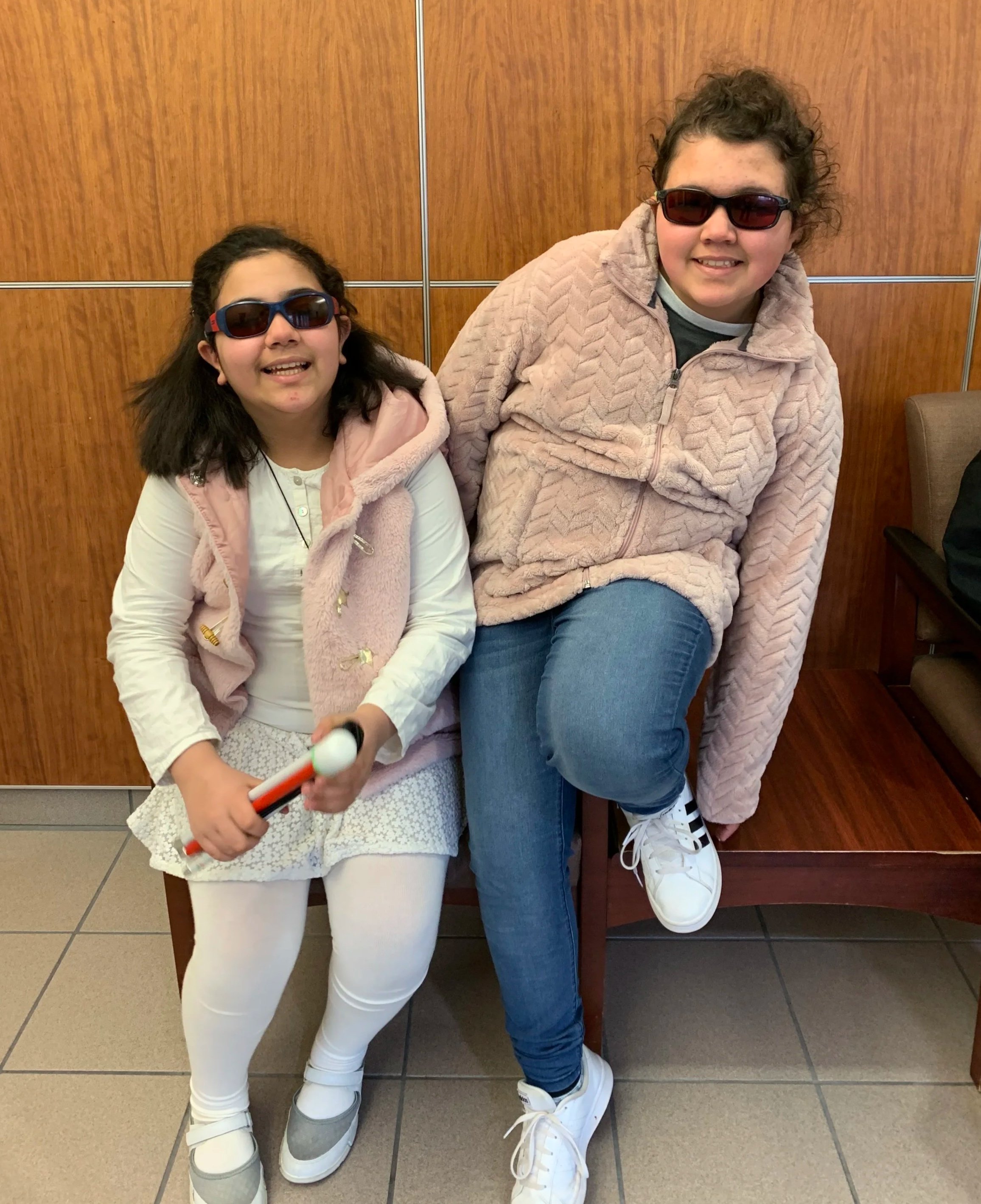 Two girls wearing sunglasses sitting on a bench, smiling. One girl is dressed in a white outfit. The other girl is dressed in a pink jacket and blue jeans.