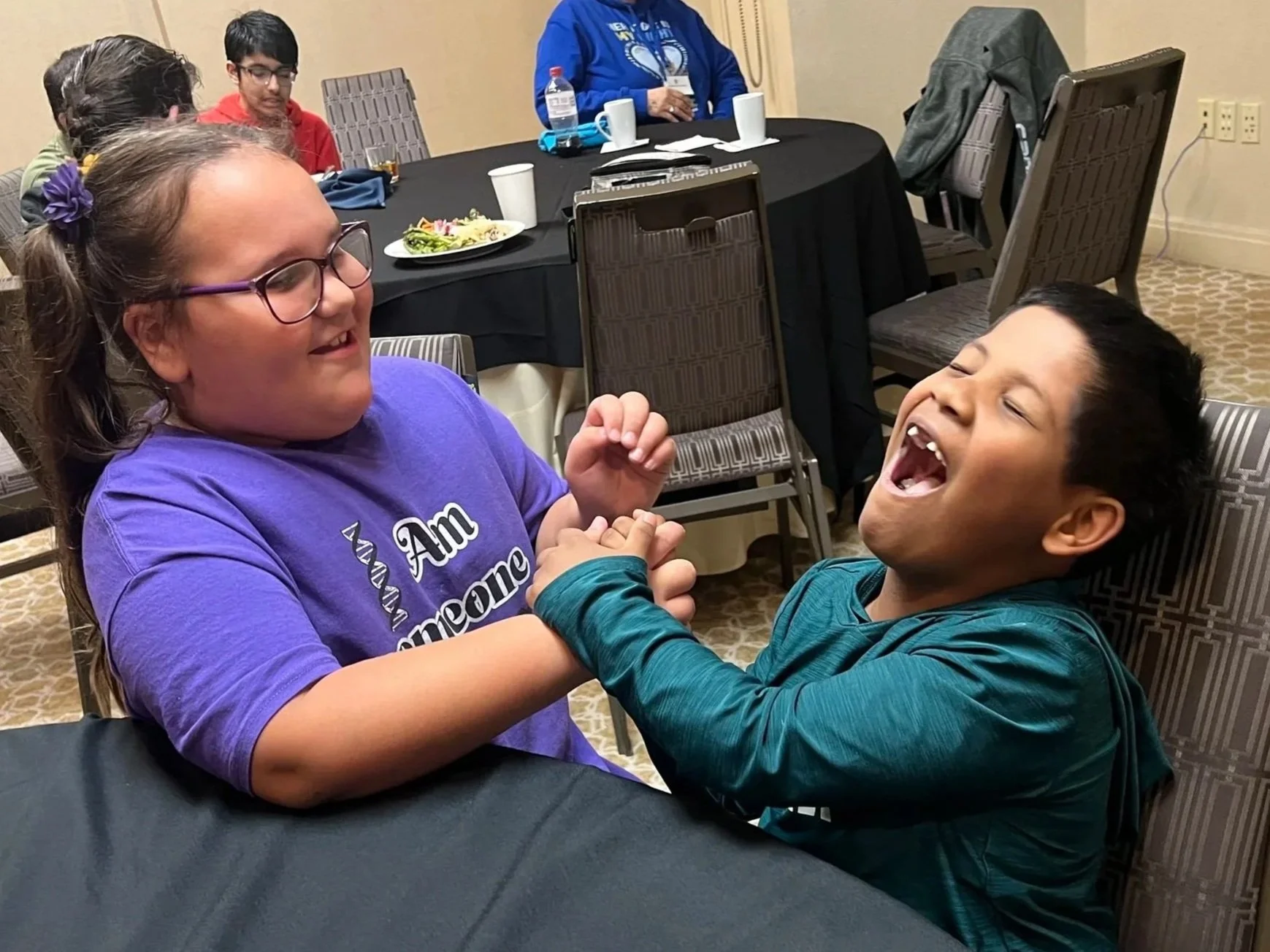 Two children, a girl with glasses and a boy, are laughing and holding hands at a table during a social gathering.