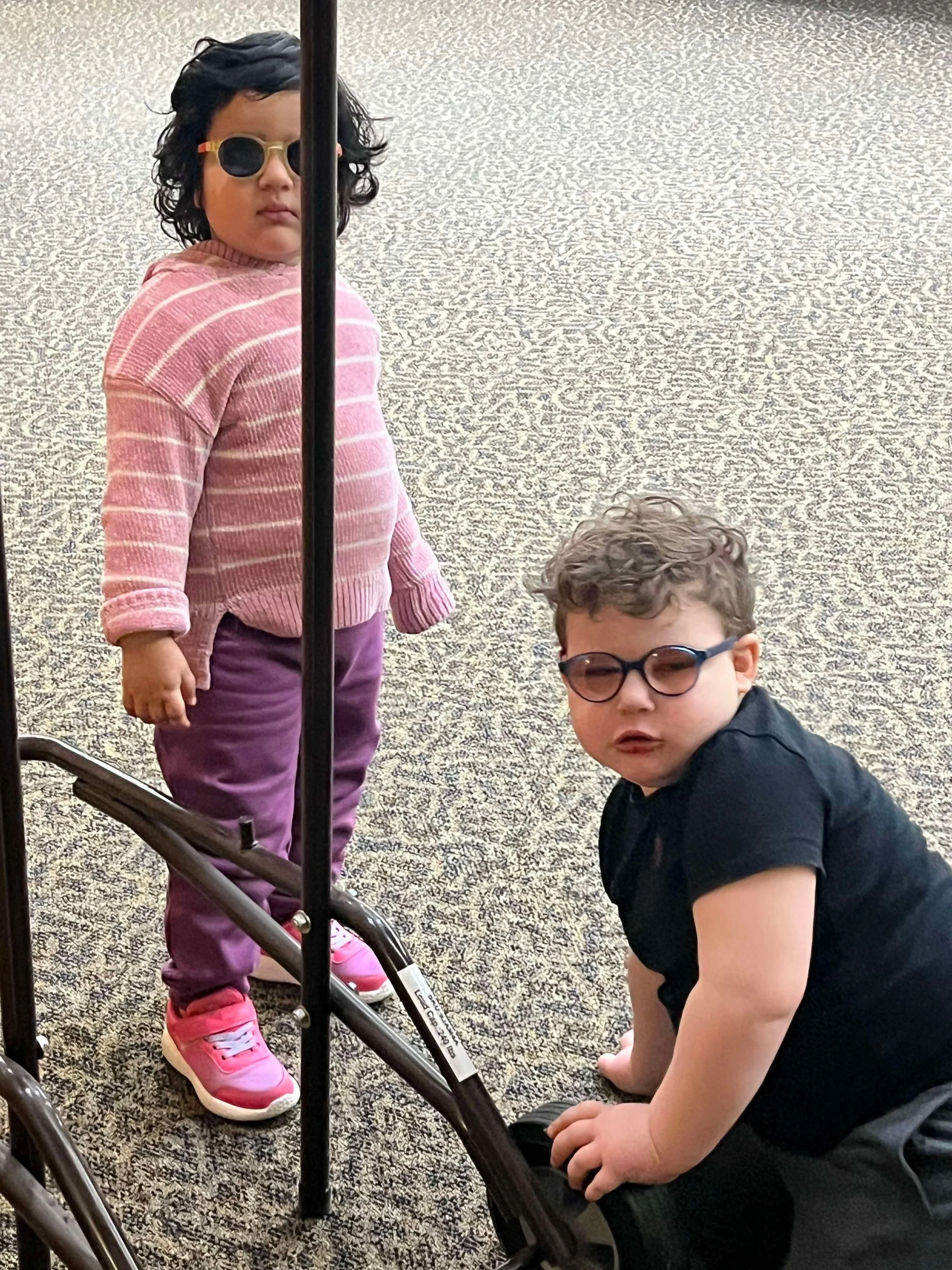 Two children, one girl and one boy, with glasses, on a carpeted floor, with a black walker in front of them.