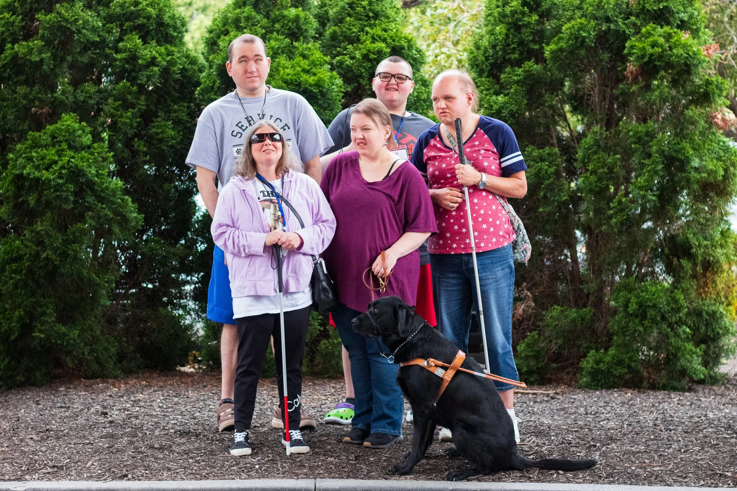 A diverse group of six people, five women and one man, posing outdoors with a black guide dog in front of green bushes. They appear to be at a park or garden during daytime.