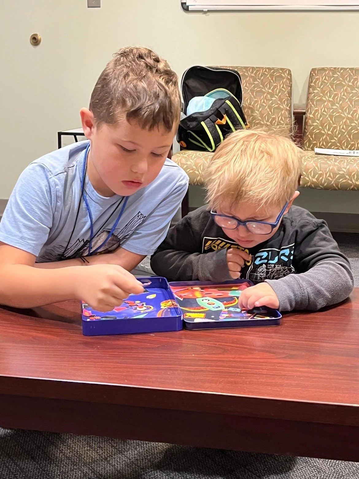 Two boys are playing a game with colorful pieces on a wooden table in a waiting room.