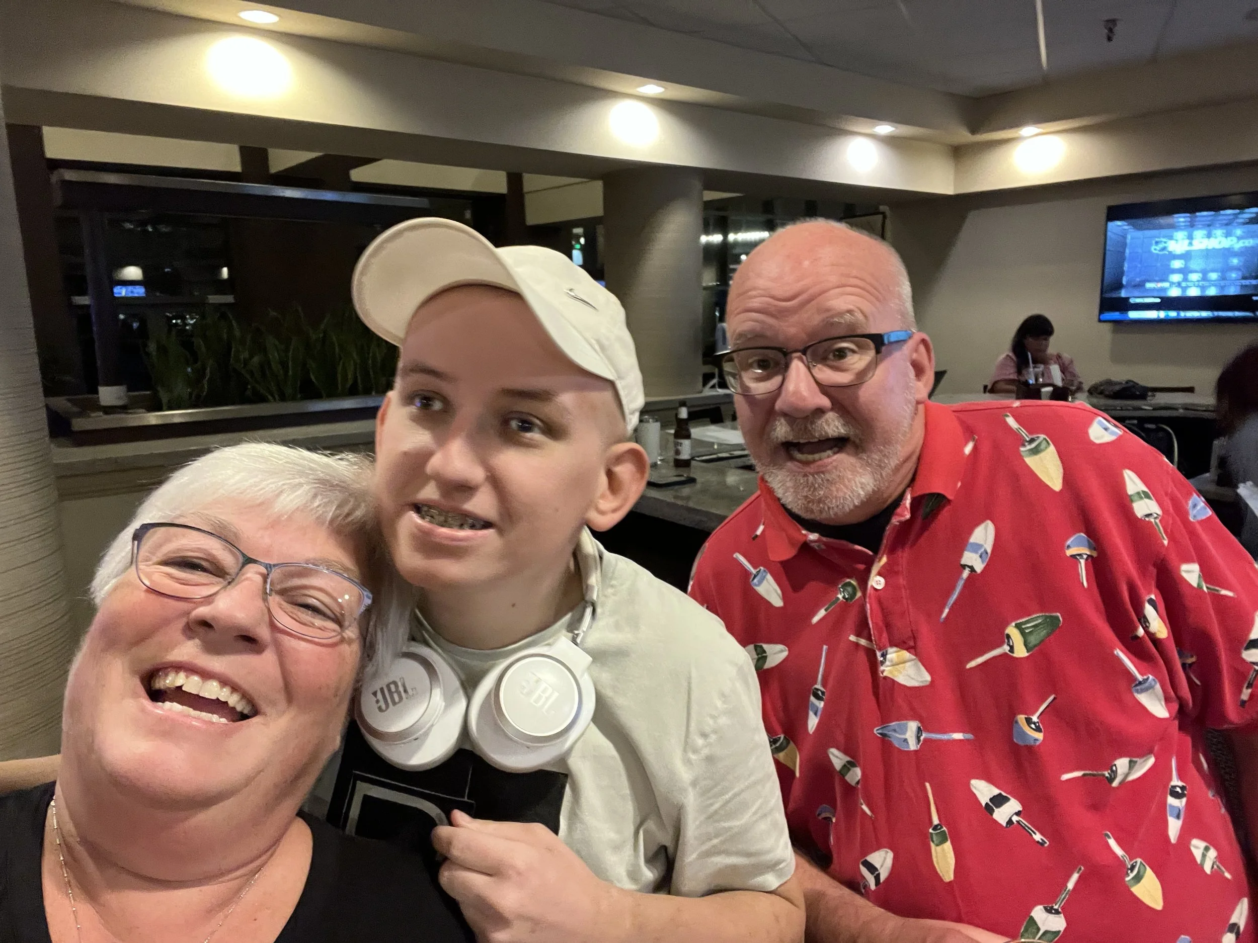 Three people smiling and posing for a photo inside a restaurant or cafe. The group includes an older woman with glasses, a young man with a white cap and headphones, and a man with glasses wearing a red shirt with a bowling pin pattern.