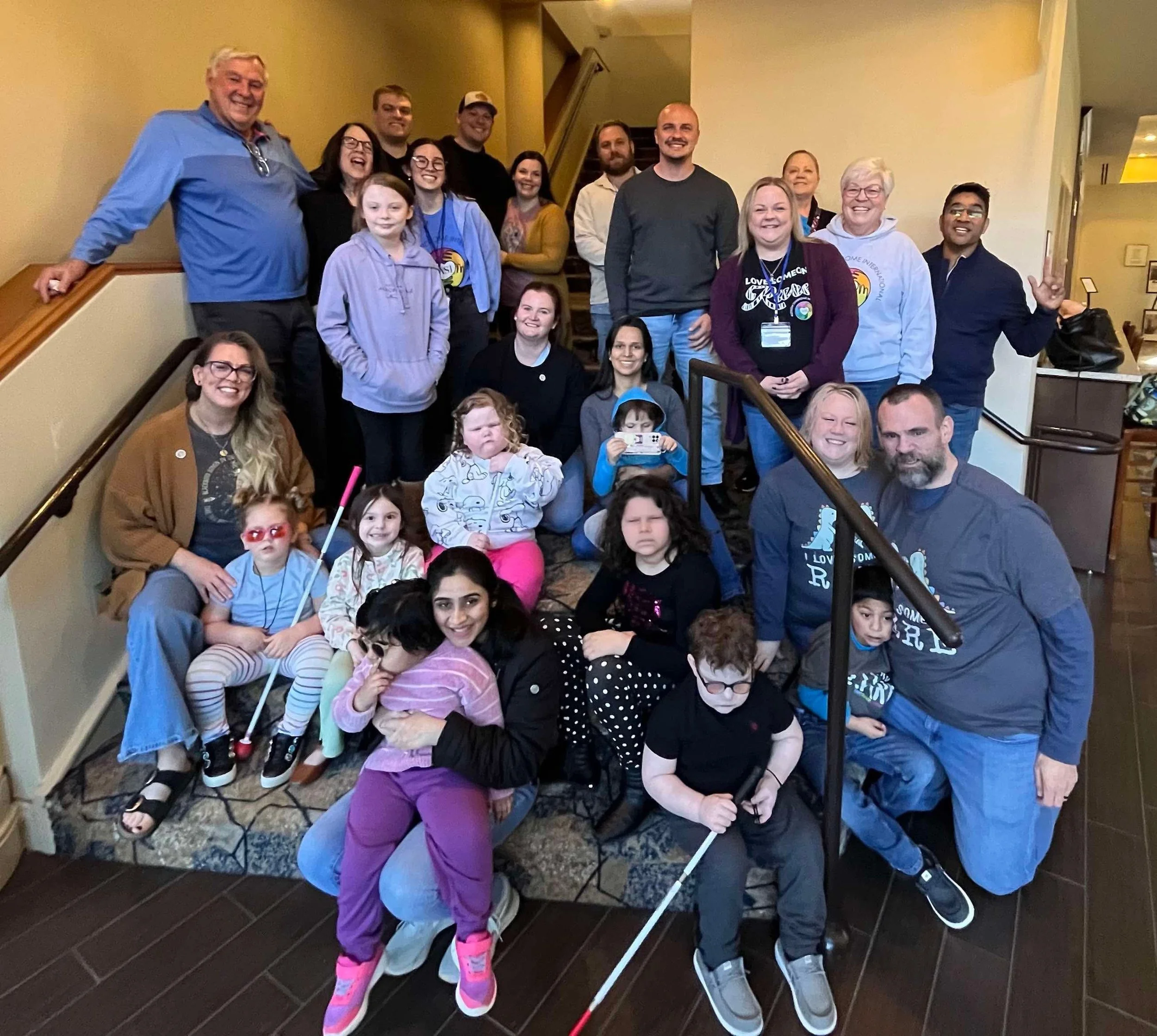 A large group of people, including children and adults, gathered on a staircase inside a building. Some children have physical disabilities and use white canes. The group is smiling and posing for a photo.