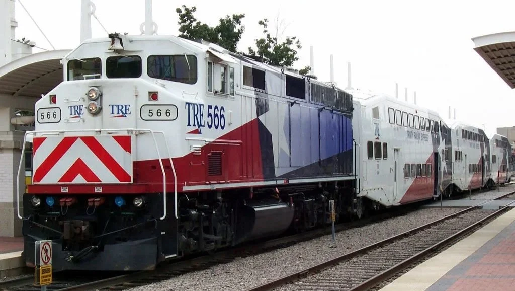 A train with the number 566 and TRE logo parked at a station, with a double-decker passenger car behind it.