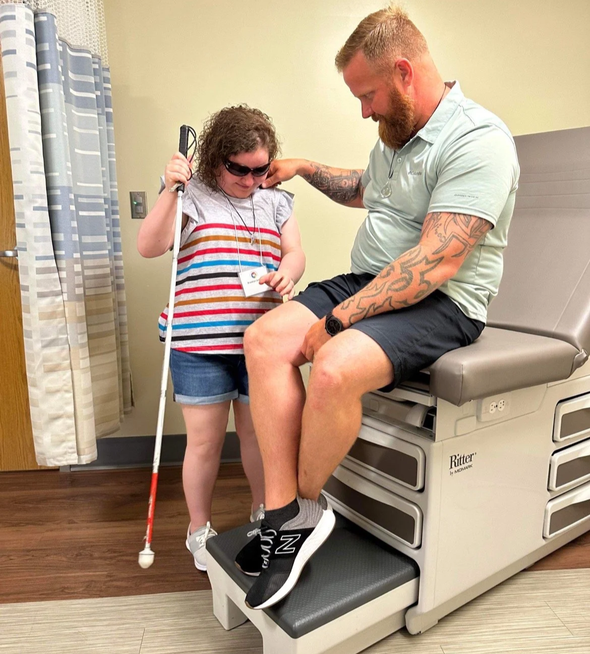 A young girl with sunglasses holding a cane and a man with tattoos sitting on an exam table in a medical room, engaging in a friendly interaction.