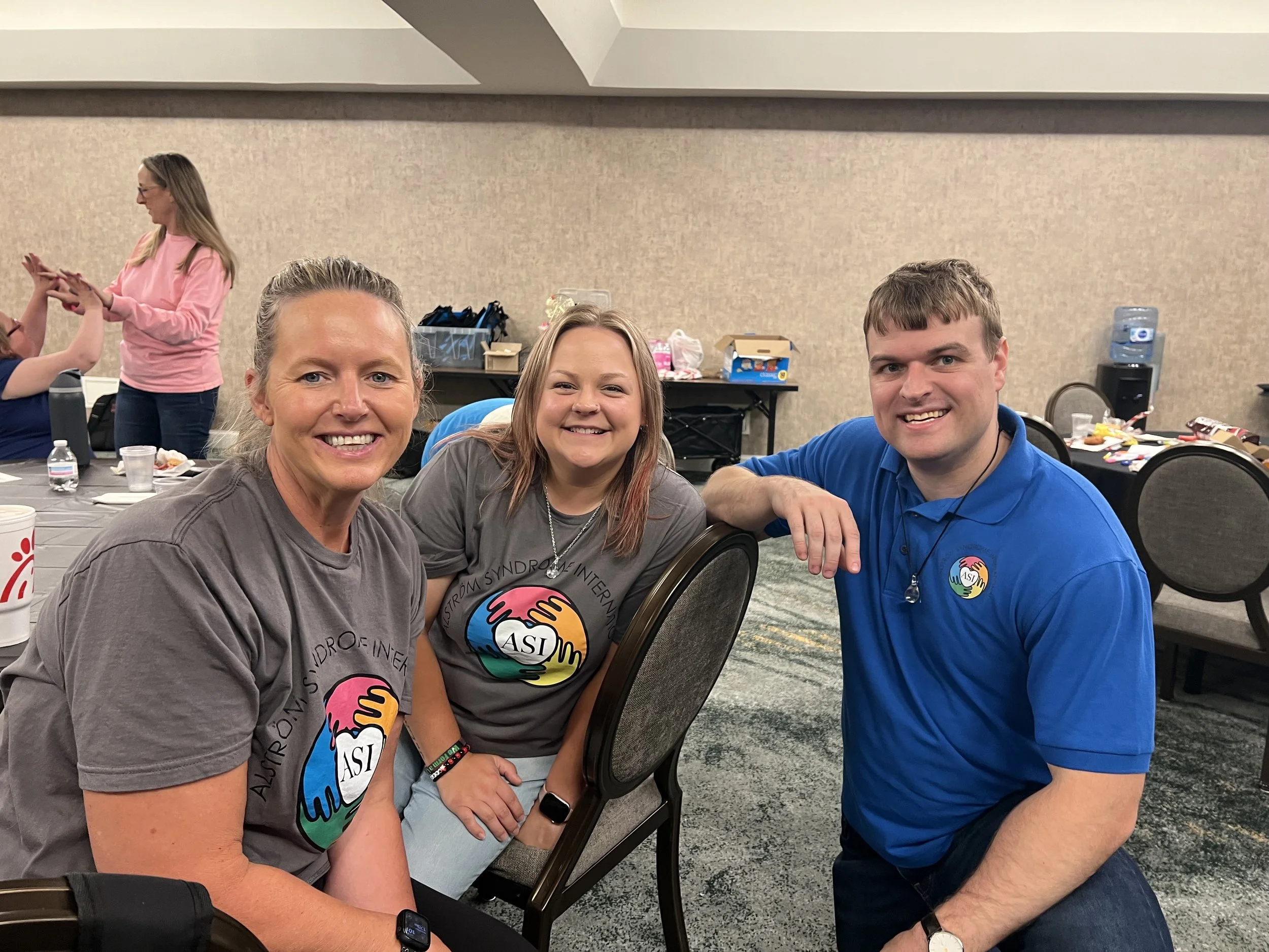Three people smiling at a table at an indoor event, with two women to the left and a man to the right, all wearing shirts with a colorful logo and the acronym 'ASI'.