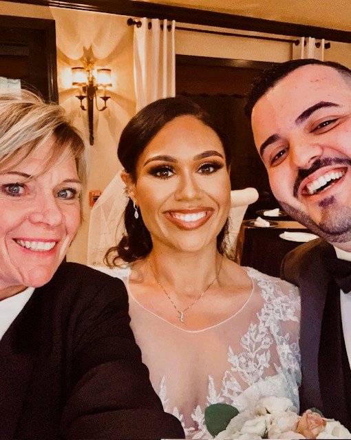 Three people smiling at a wedding reception, including a bride in a white wedding dress and two guests, one woman and one man.