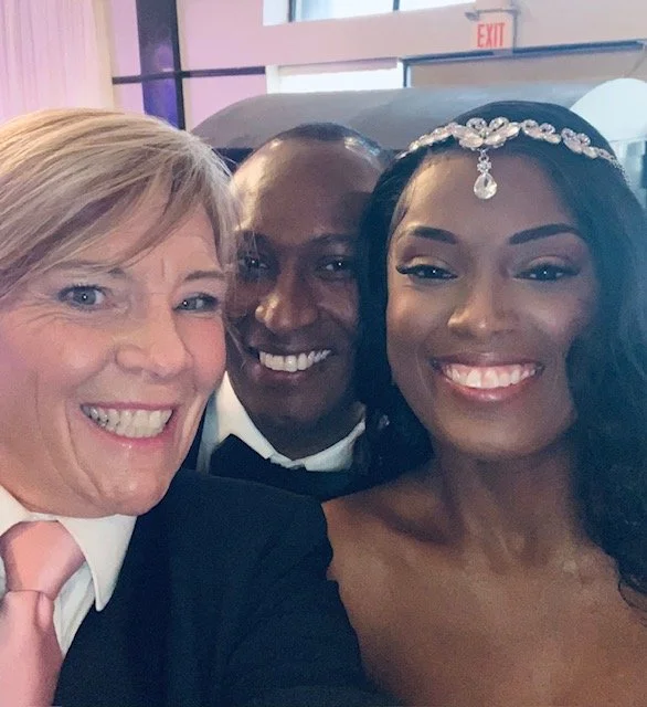 Three smiling people taking a selfie at a wedding reception, with the bride wearing a tiara and the groom in formal attire.