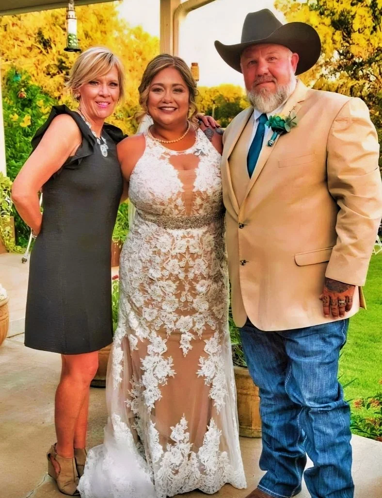Three people, two women and one man, posing outdoors at a wedding. The woman in the middle is wearing a lace wedding dress, and the woman on the left is wearing a dark dress. The man is dressed in a light-colored suit with a cowboy hat. All are smiling in a garden setting with yellow flowers and trees in the background.