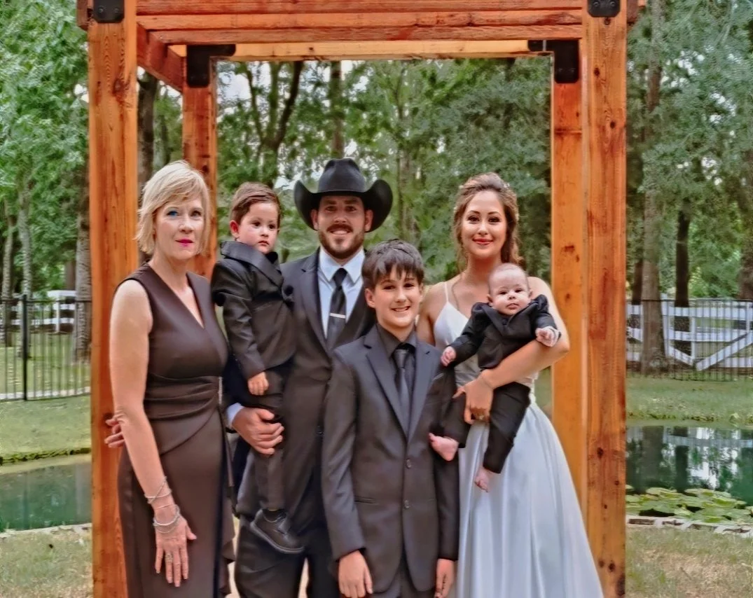 A group of six people, including two children, standing under a wooden archway outdoors in a park-like setting with trees and a pond in the background, all dressed formally or in special attire.