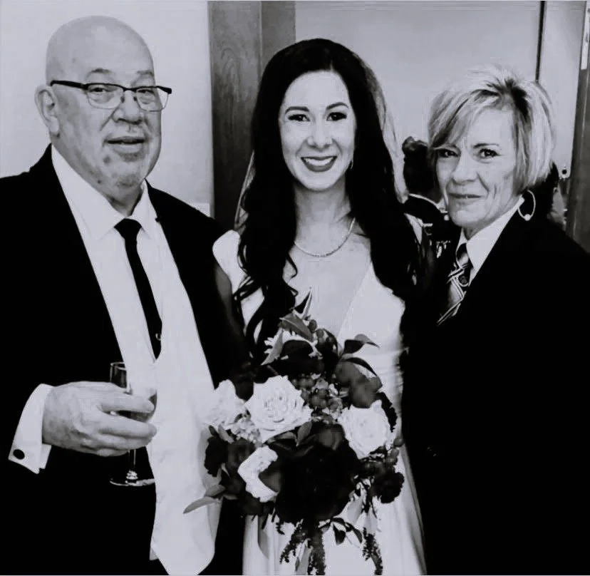 Three people at a formal event, with one holding a bouquet of flowers and a glass of wine, dressed in suits and a dress, smiling in front of a wooden background.