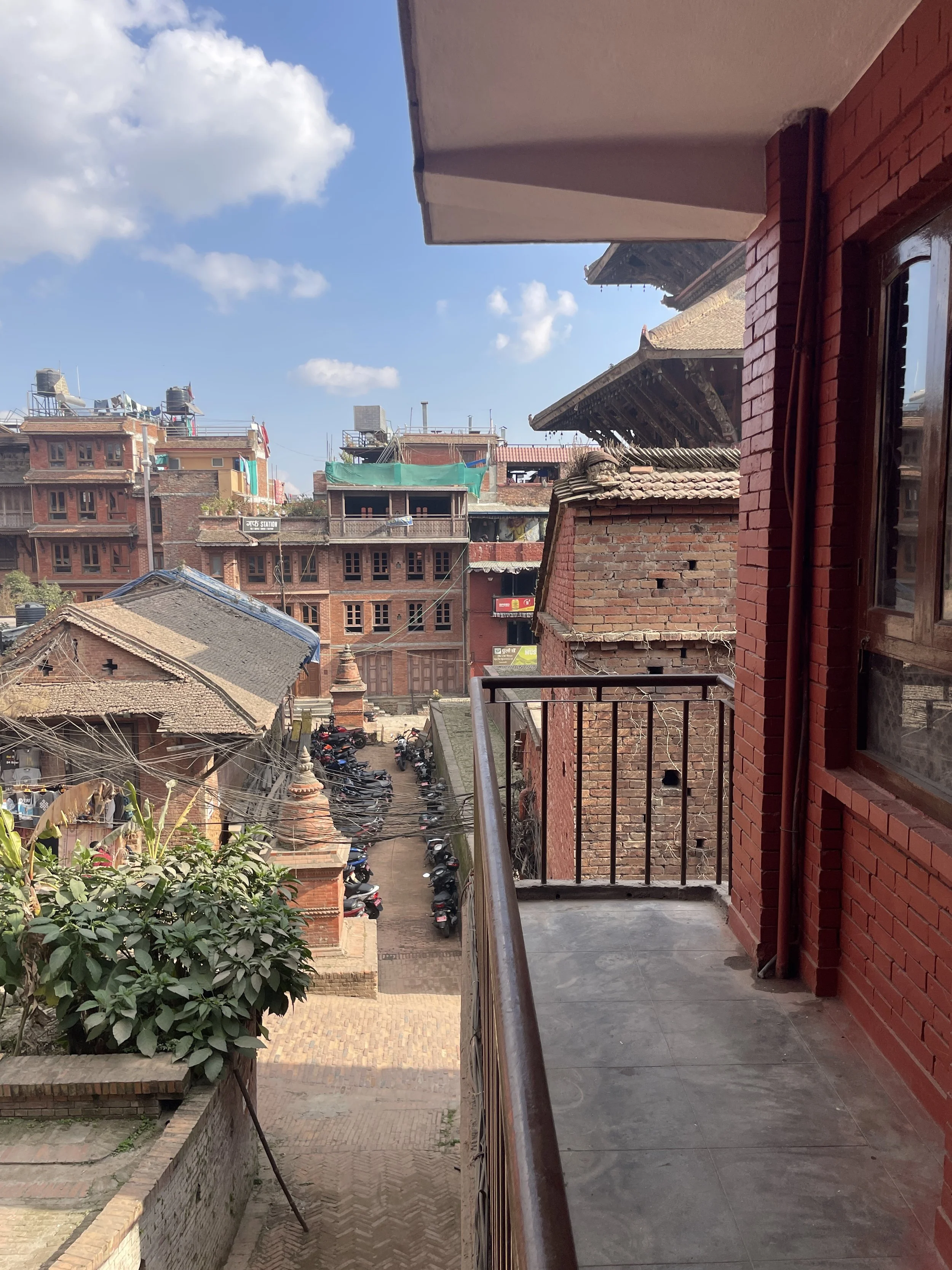 View from a balcony showing traditional brick buildings, a narrow street with parked motorcycles, and a partly cloudy blue sky.