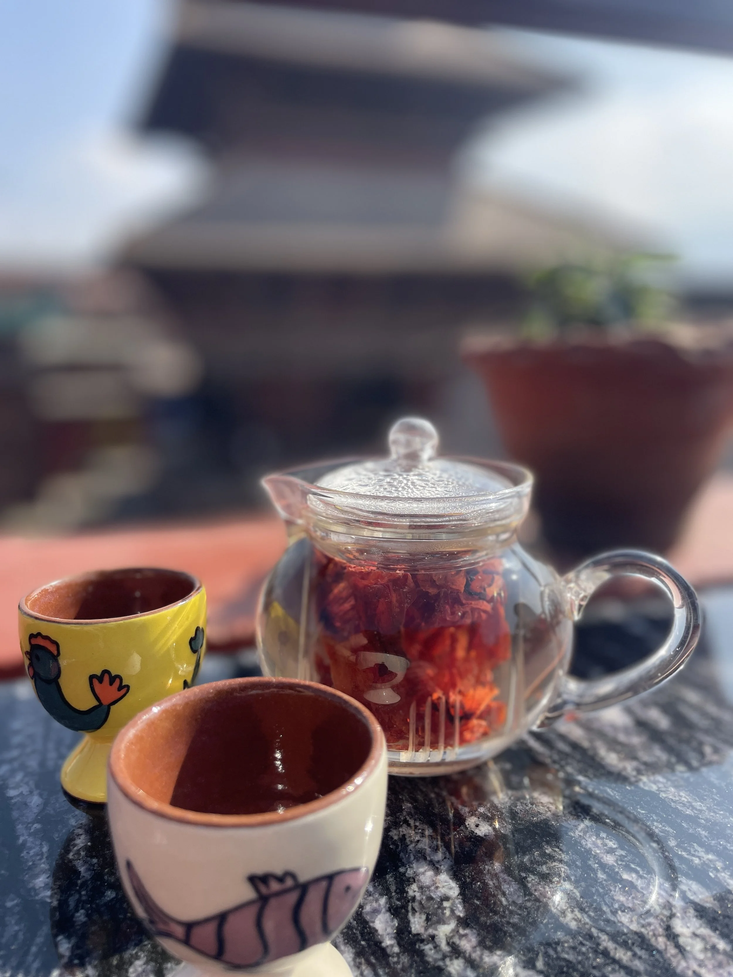 A glass teapot filled with dried petals rests on the rooftop table, paired with handmade ceramic cups. In the background, Nyatapola Temple rises softly into view, grounding the moment in place.
