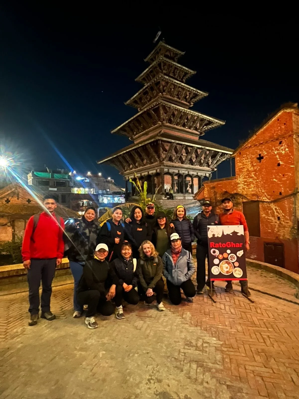 A group of people posing in front of the 5-story pagoda-style temple at night, with a sign reading 'RatoGhar'; located in an outdoor setting with brick walls and a cobblestone ground.
