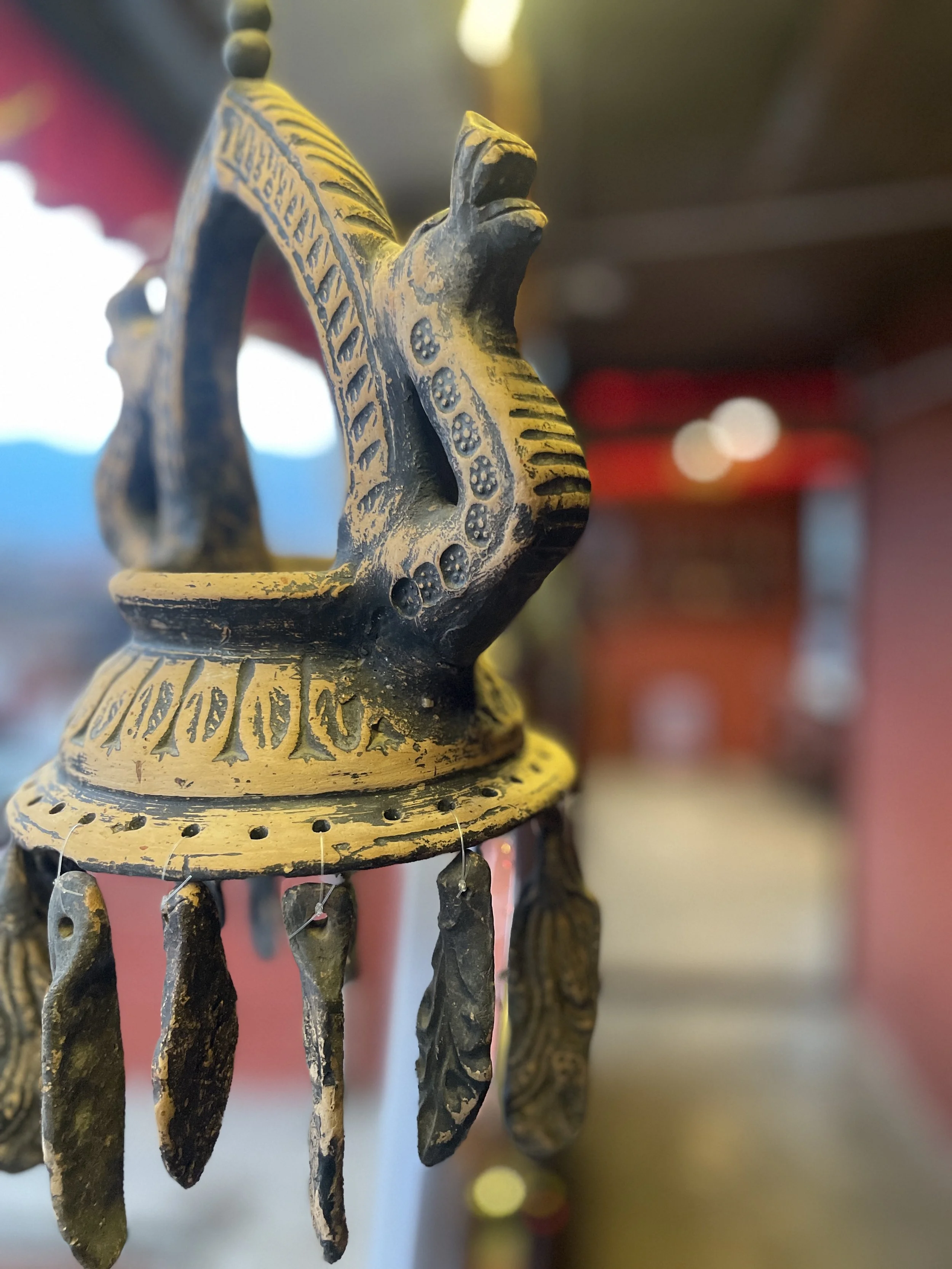 Close-up of a decorative Nepali pottery lantern, featuring hanging feathers or leaves, in a blurred indoor setting with warm lighting.