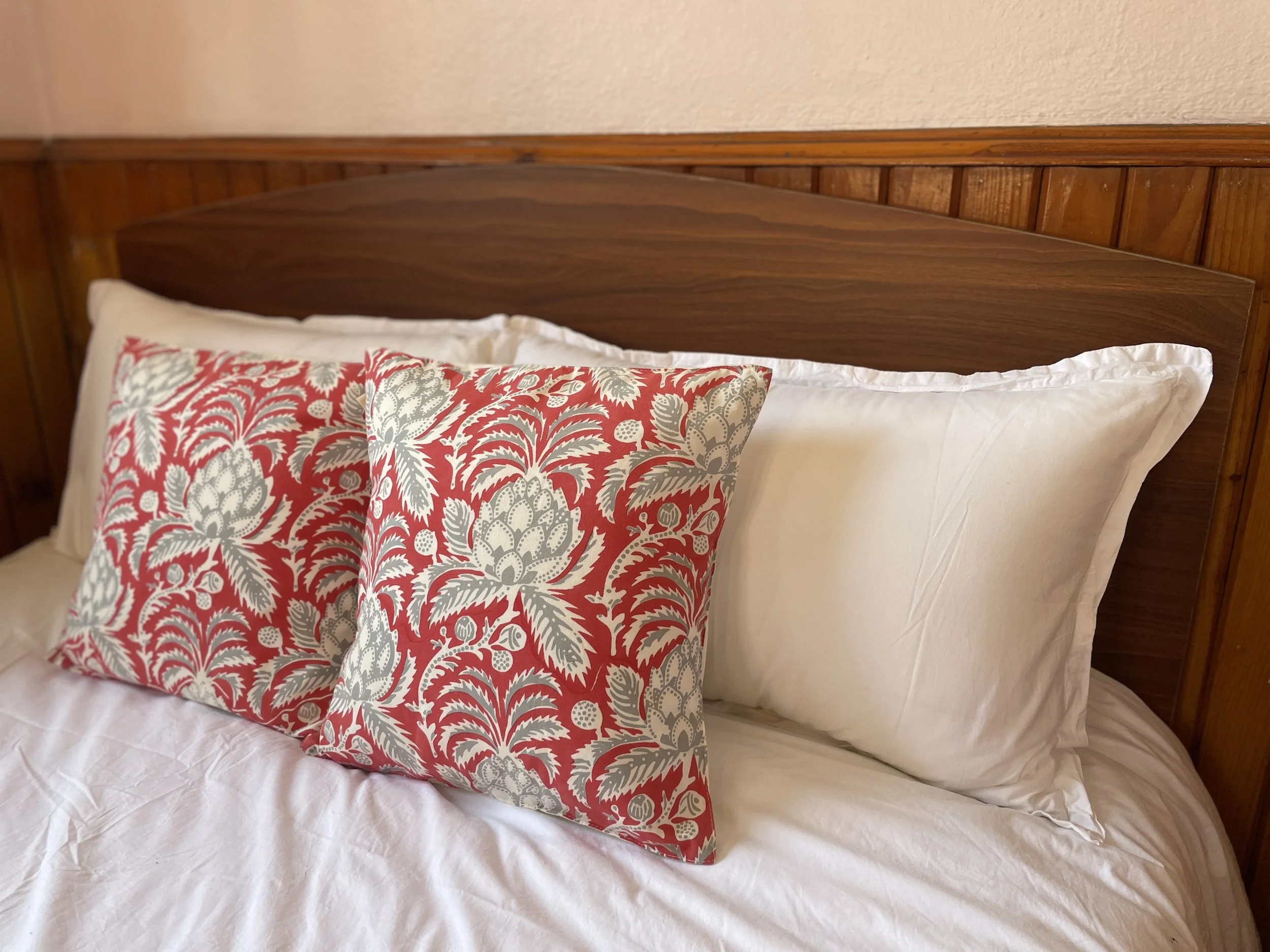 Decorative bed pillows with a floral pattern in red and gray on white, on a bed with white sheets and wooden headboard.