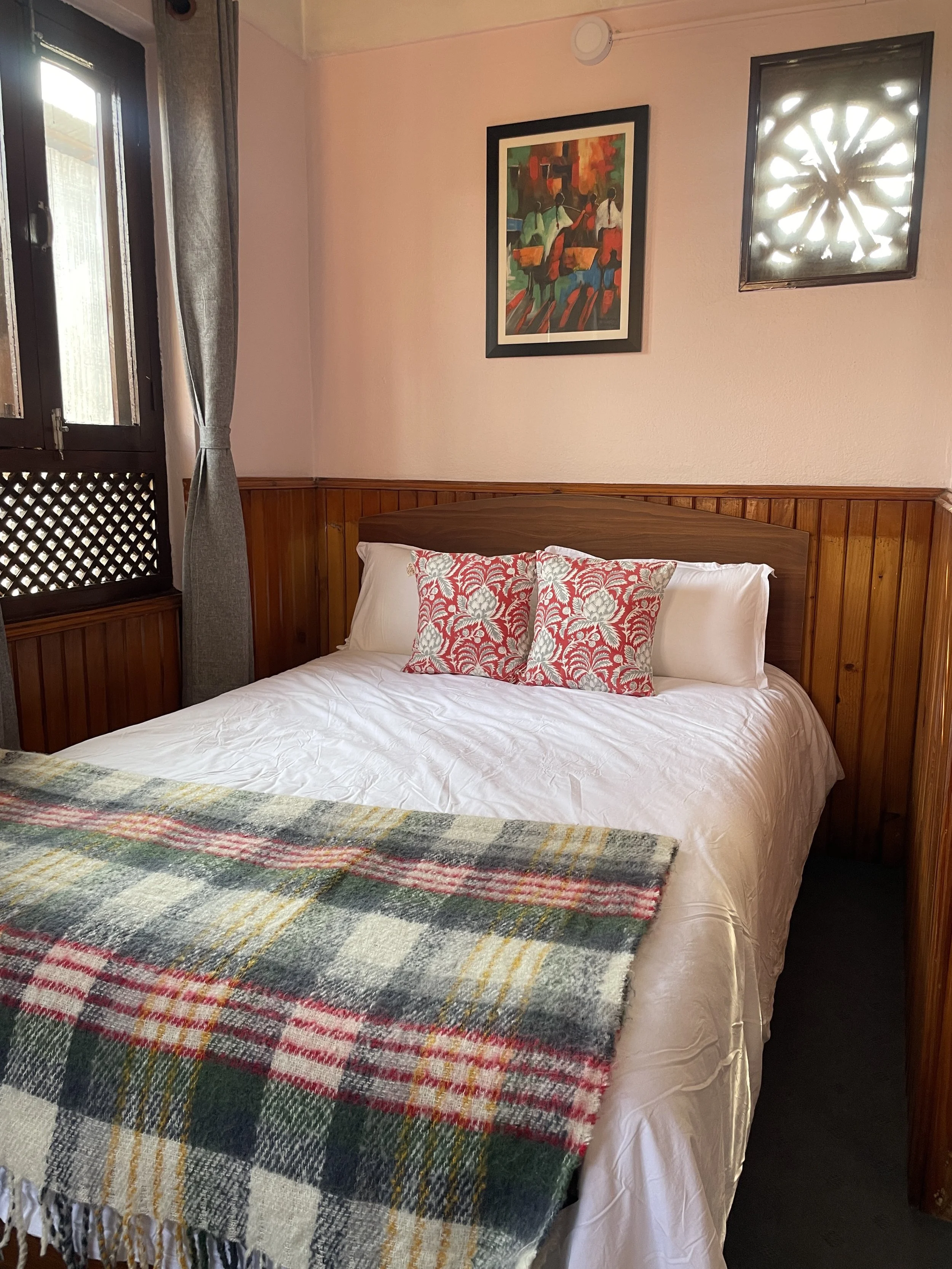 A cozy bedroom with a bed featuring white sheets, two white pillows, and three decorative red and white patterned pillows. There is a plaid blanket with green, red, yellow, and white on the foot of the bed. The room has a wooden headboard, wood-panel