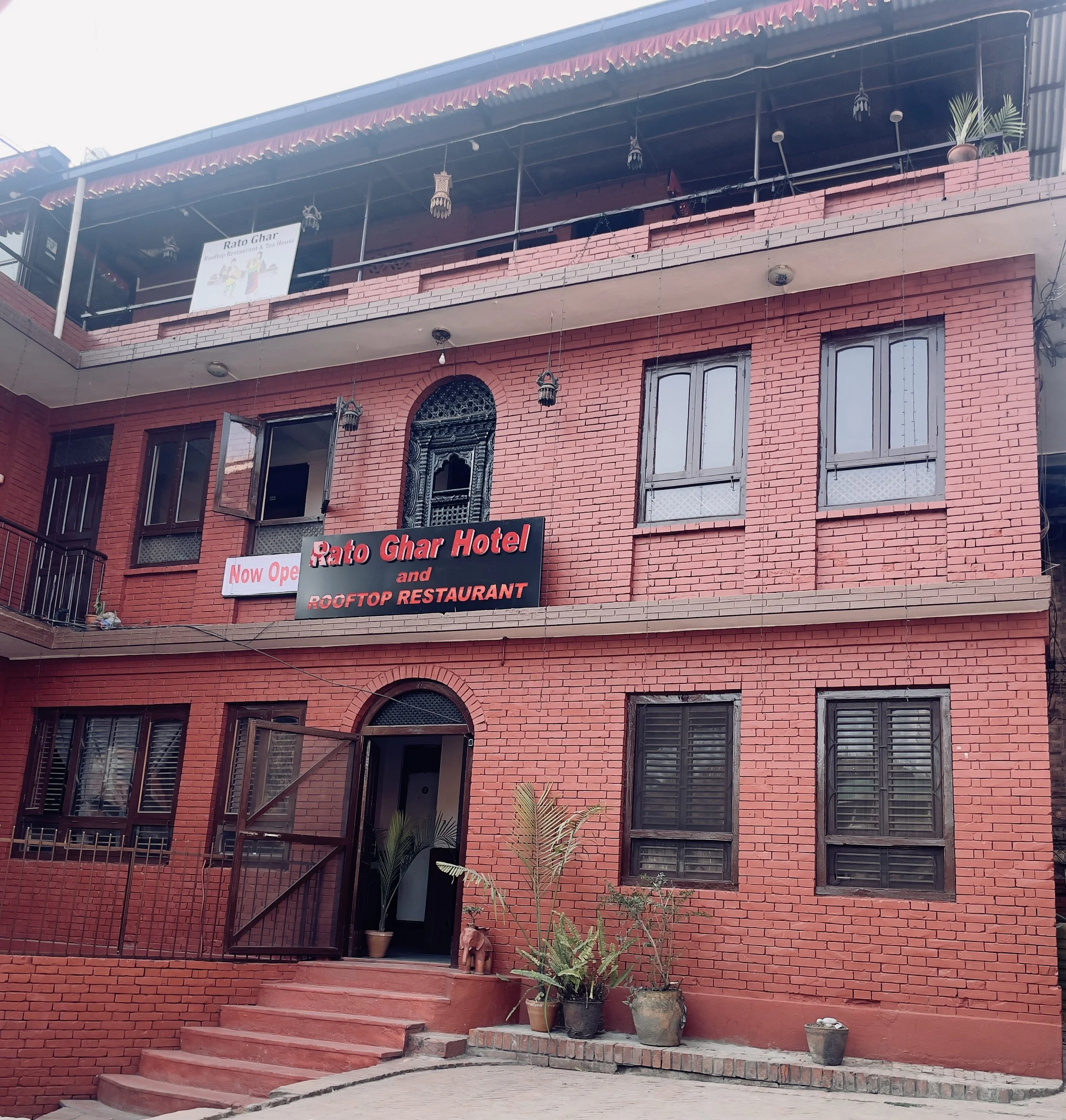 Red brick building with signs introducing Rato Ghar Hotel and Rooftop Restaurant, featuring multiple windows, a gate, stairs, and potted plants outside.