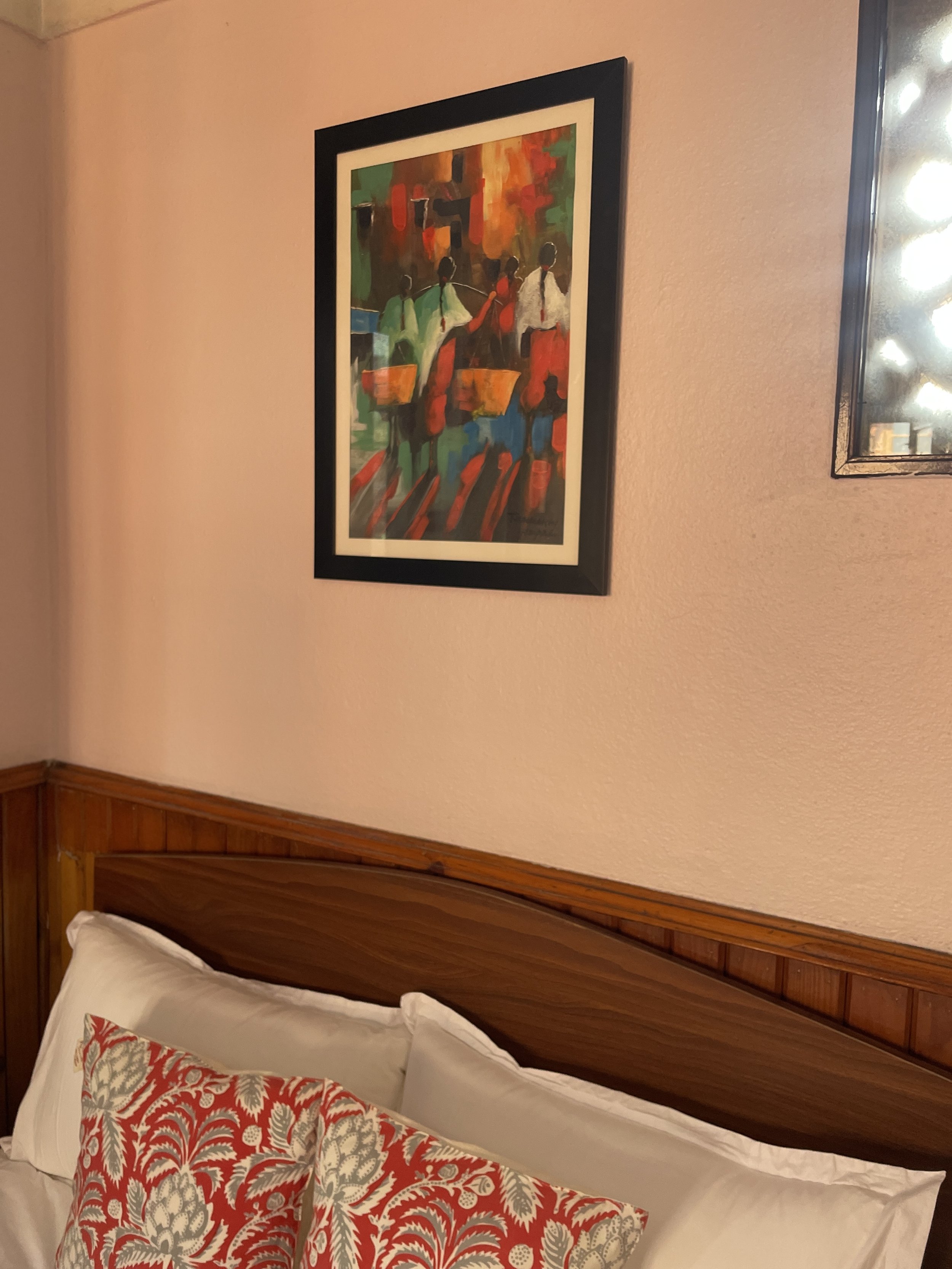 A framed colorful painting of three figures standing outdoors, hung on a light pink wall above a wooden headboard with white and patterned pillows on a bed.