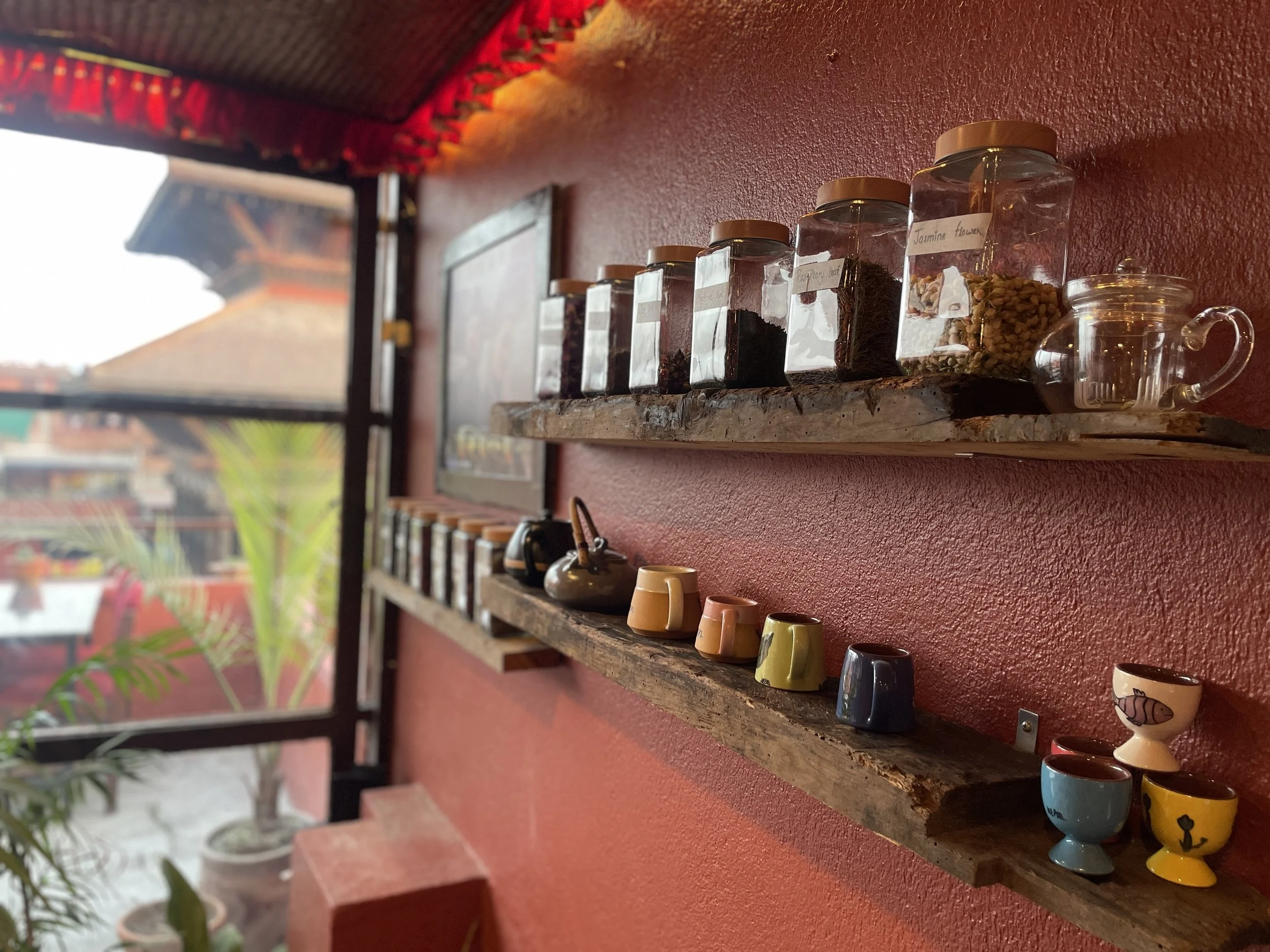 Two wooden shelves mounted on a red wall display glass jars filled with various loose-leaf teas, with colorful cups and a black teapot on the lower shelf, near a window with a view of a brick pagoda style temple and potted plants outside.