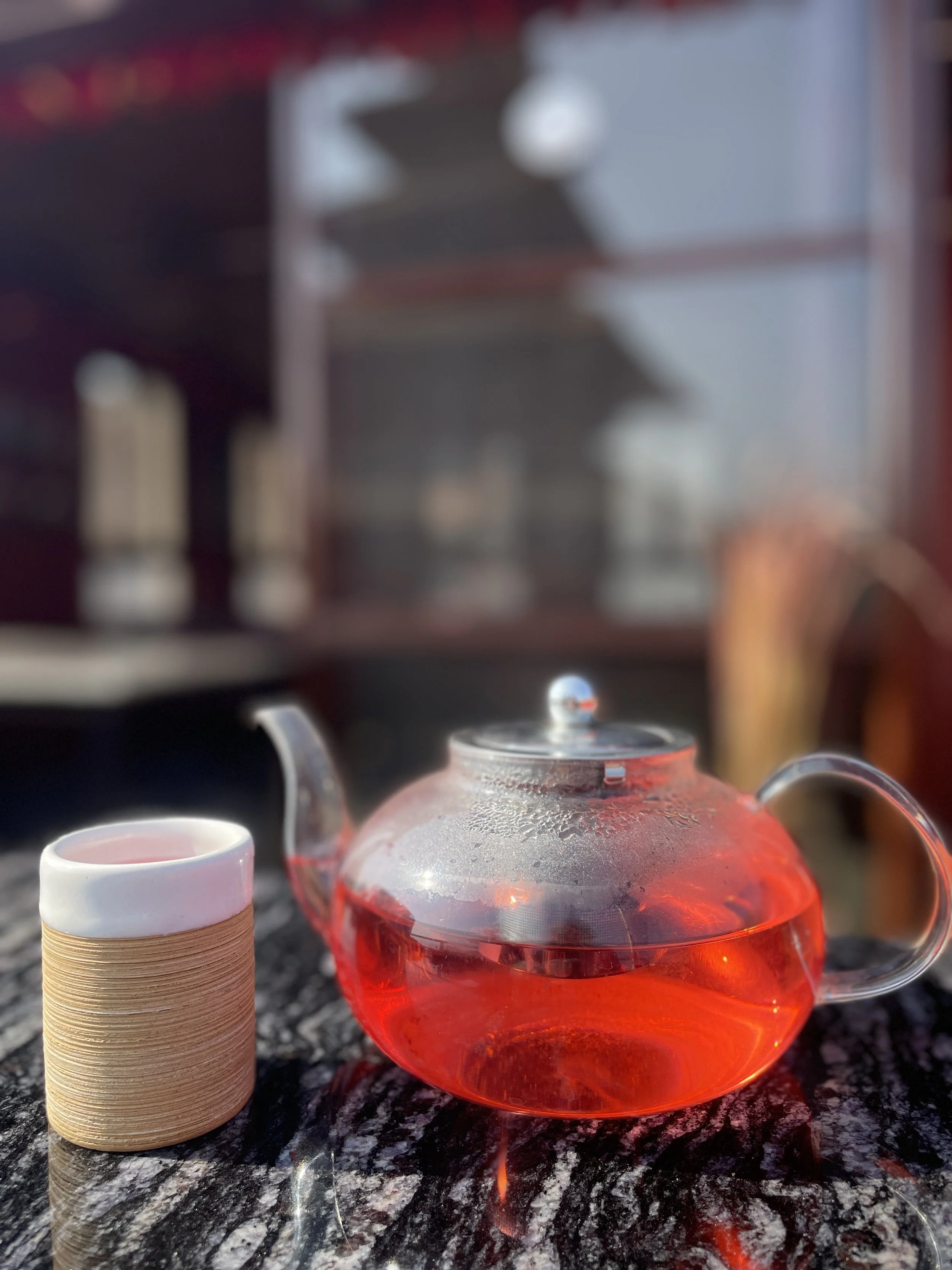 A glass teapot filled with red tea and a small ceramic cup on a dark marble table outside, with a blurred background of a building or structure.