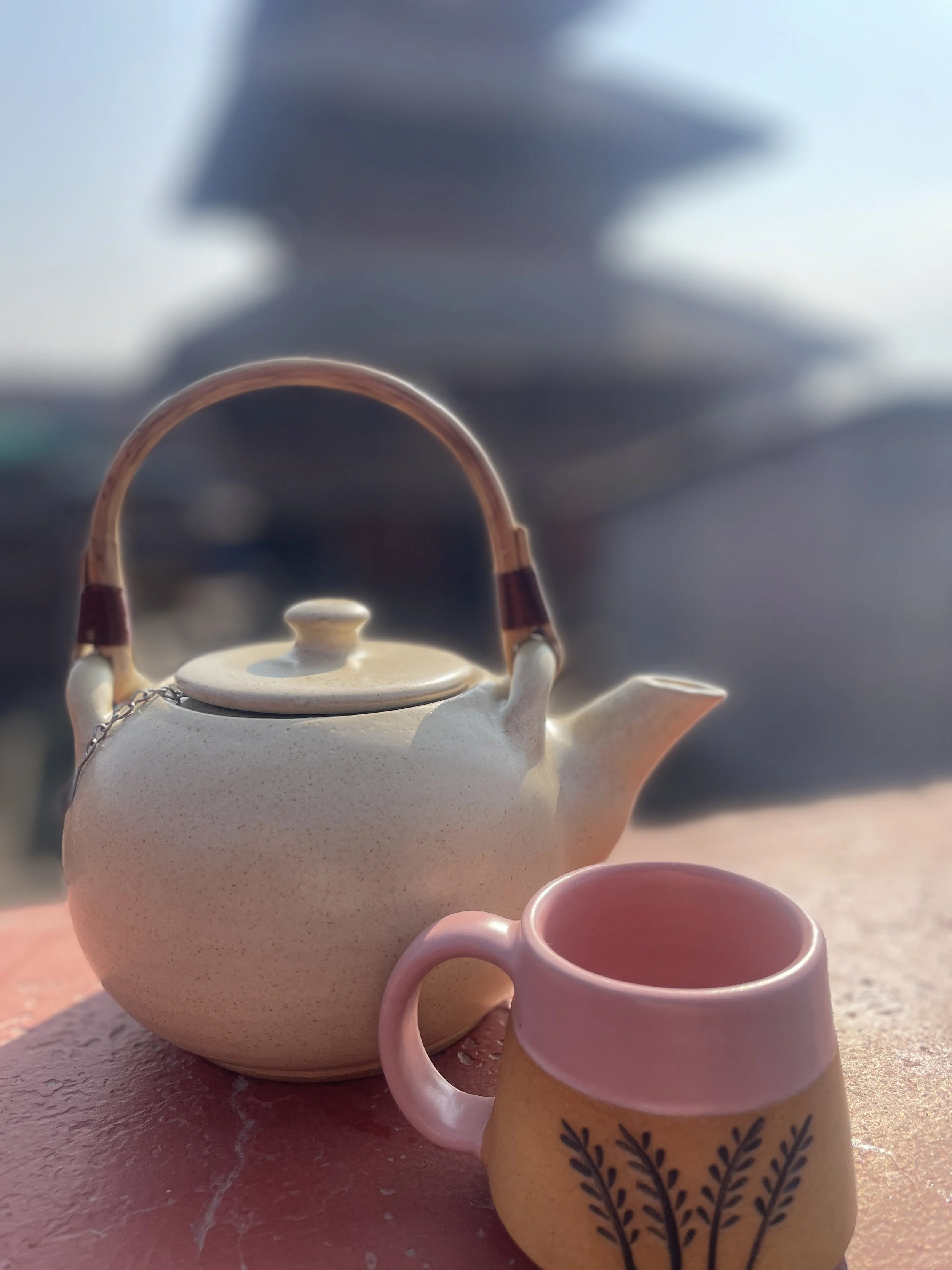 A ceramic teapot with a woven handle and a matching pink mug with a decorative design on an outdoor table, with a traditional Newari pagoda building blurred in the background.