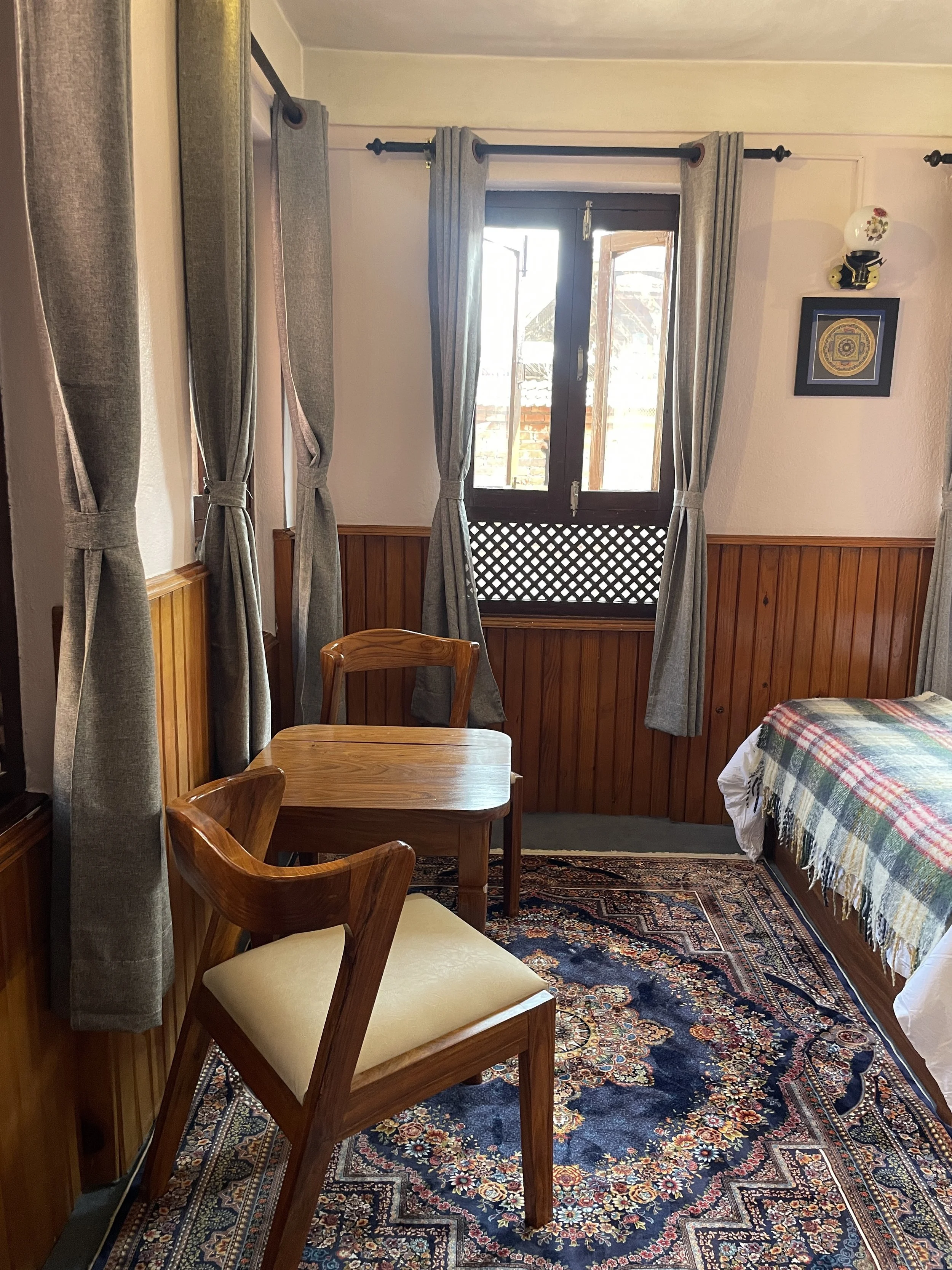 A cozy bedroom with wooden paneling on the walls, a window with gray curtains, and a small wooden table with two chairs. There is a bed with a plaid blanket, a decorative rug on the floor, and framed wall artwork.