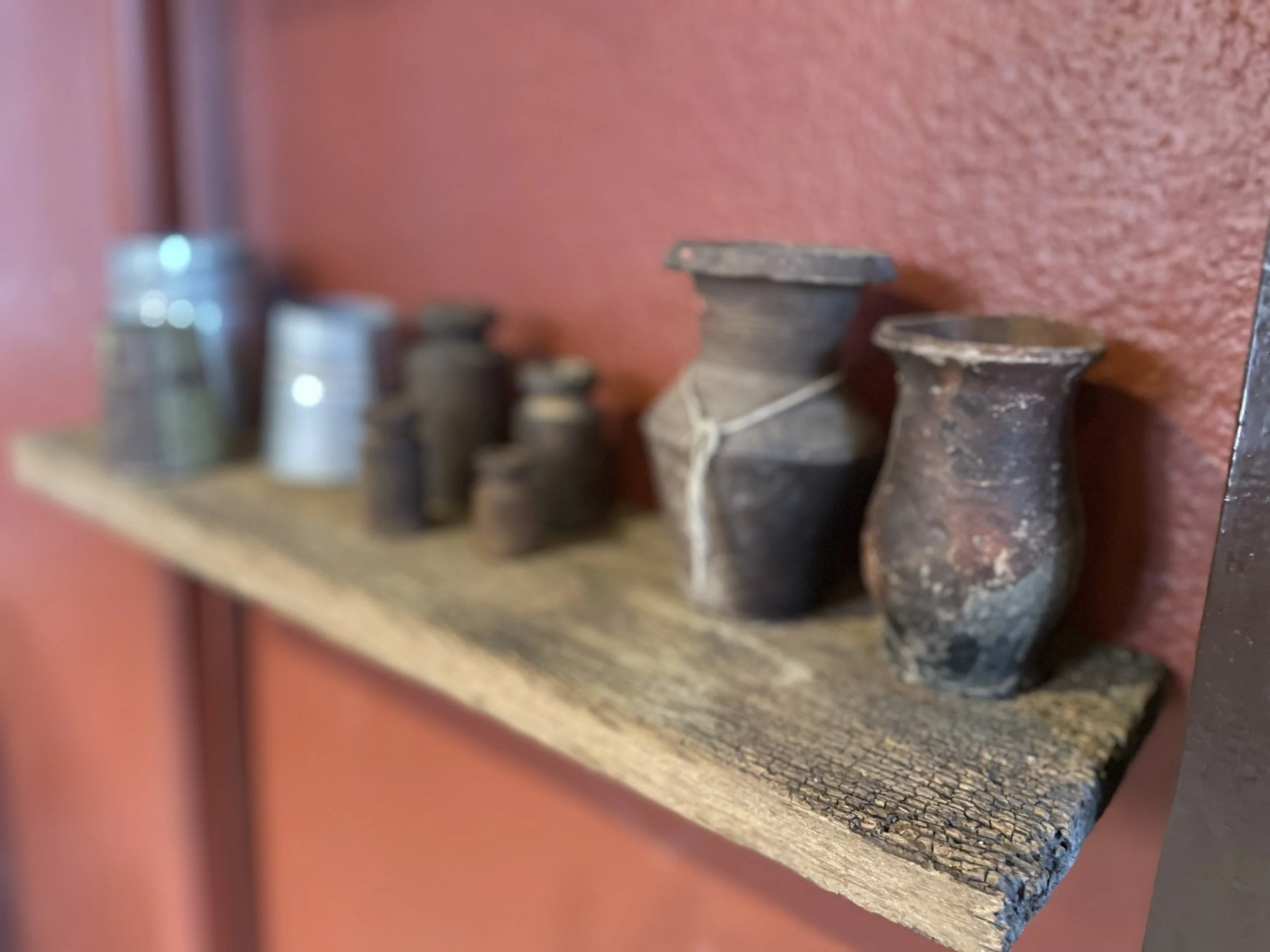 A series of small, ancient, weathered clay or ceramic vessels of varying sizes and shapes arranged on a rustic wooden shelf against a textured red wall.