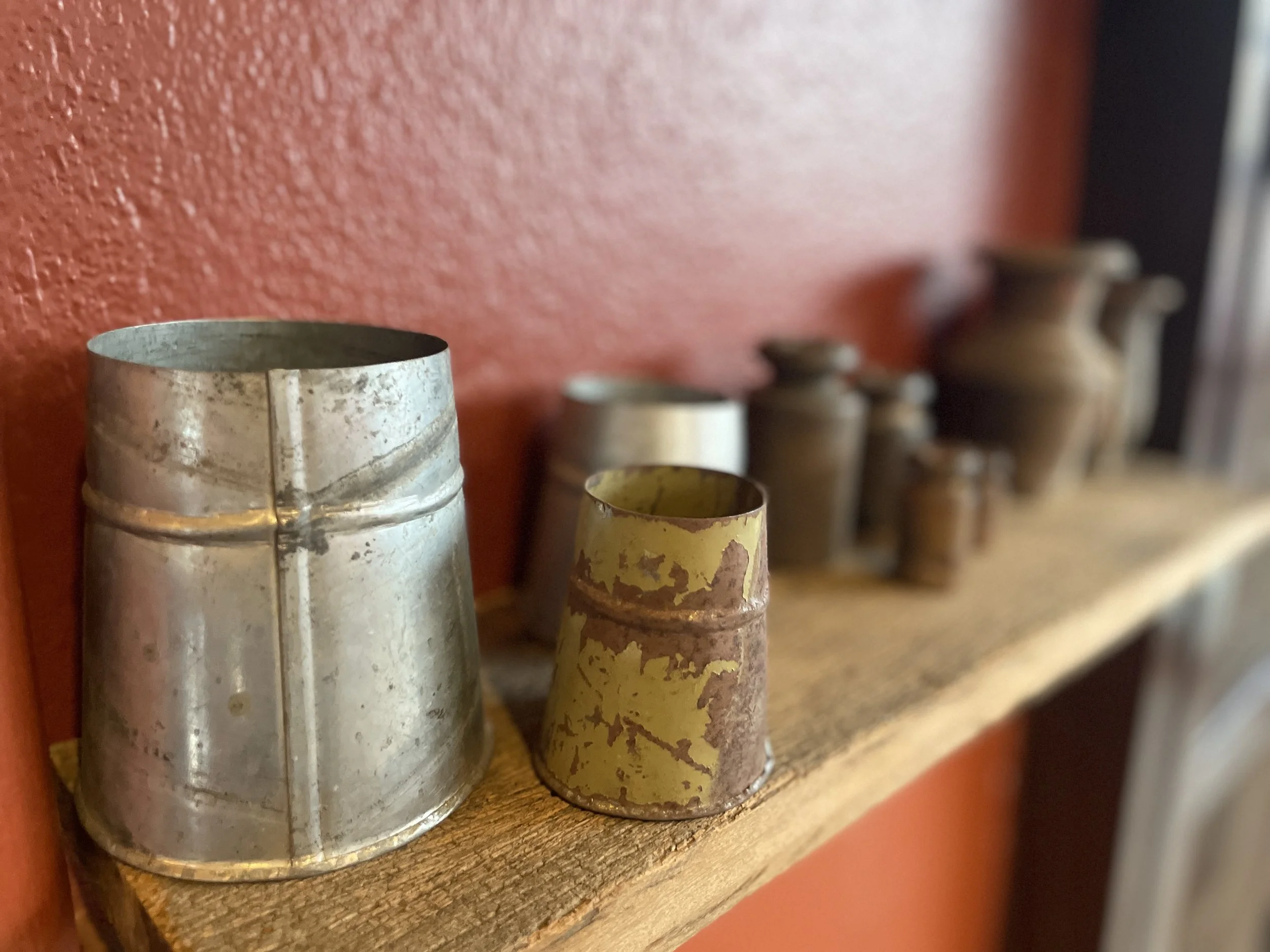 A row of old, rusty, and weathered metal containers of various sizes lined up on a wooden shelf against a textured reddish-orange wall.