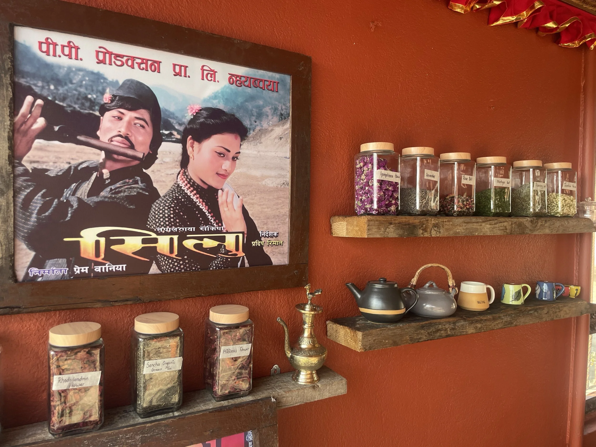 A wall decorated with vintage Nepali movie poster, jars of loose-leaf teas, a traditional teapot, cups, and saucers on rustic wooden shelves.