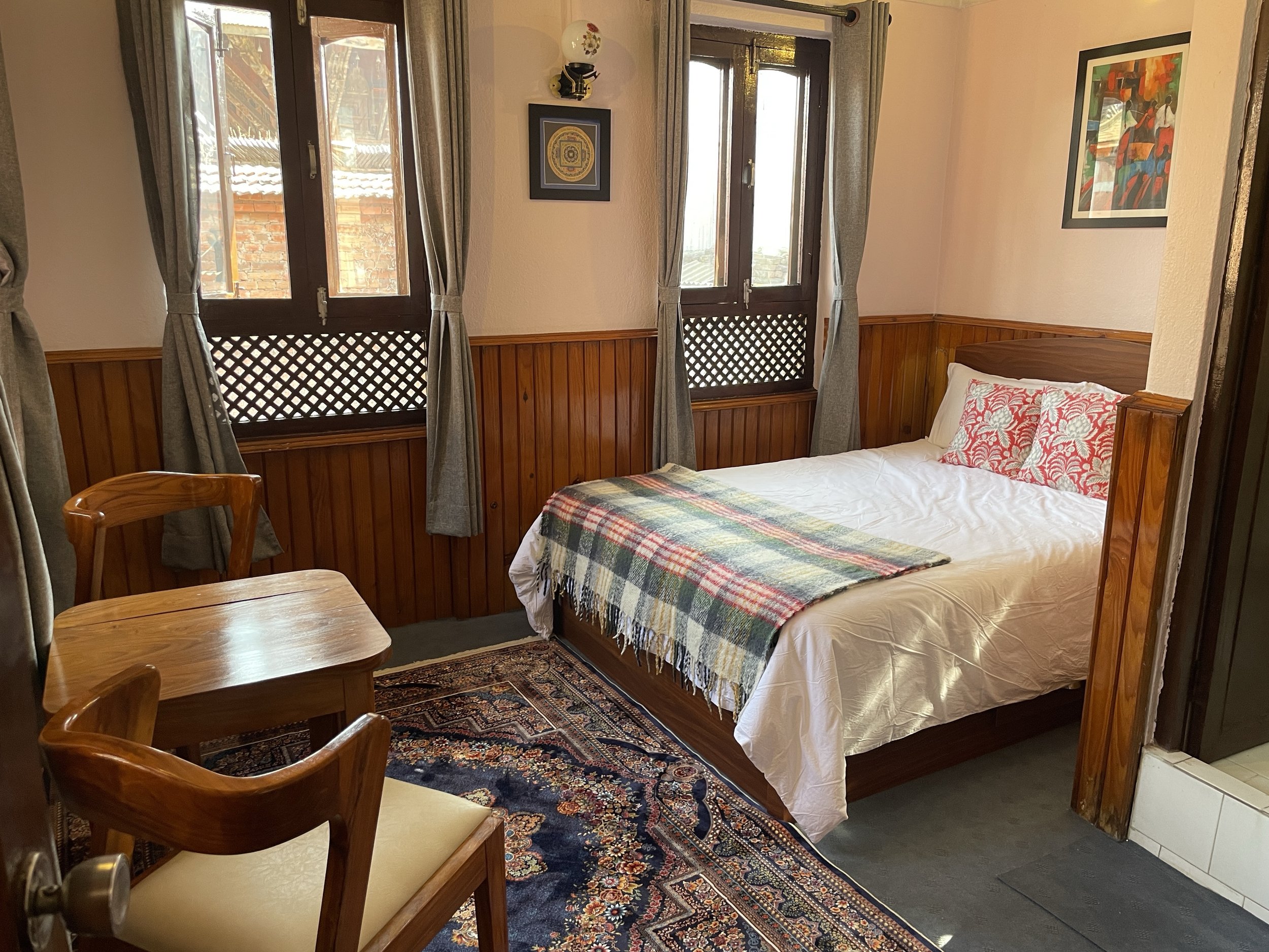 A bedroom with a bed, wooden headboard, white bedding, and red patterned pillows. There are three windows with gray curtains, a small wooden table with two chairs, a patterned rug, and wooden wall paneling.