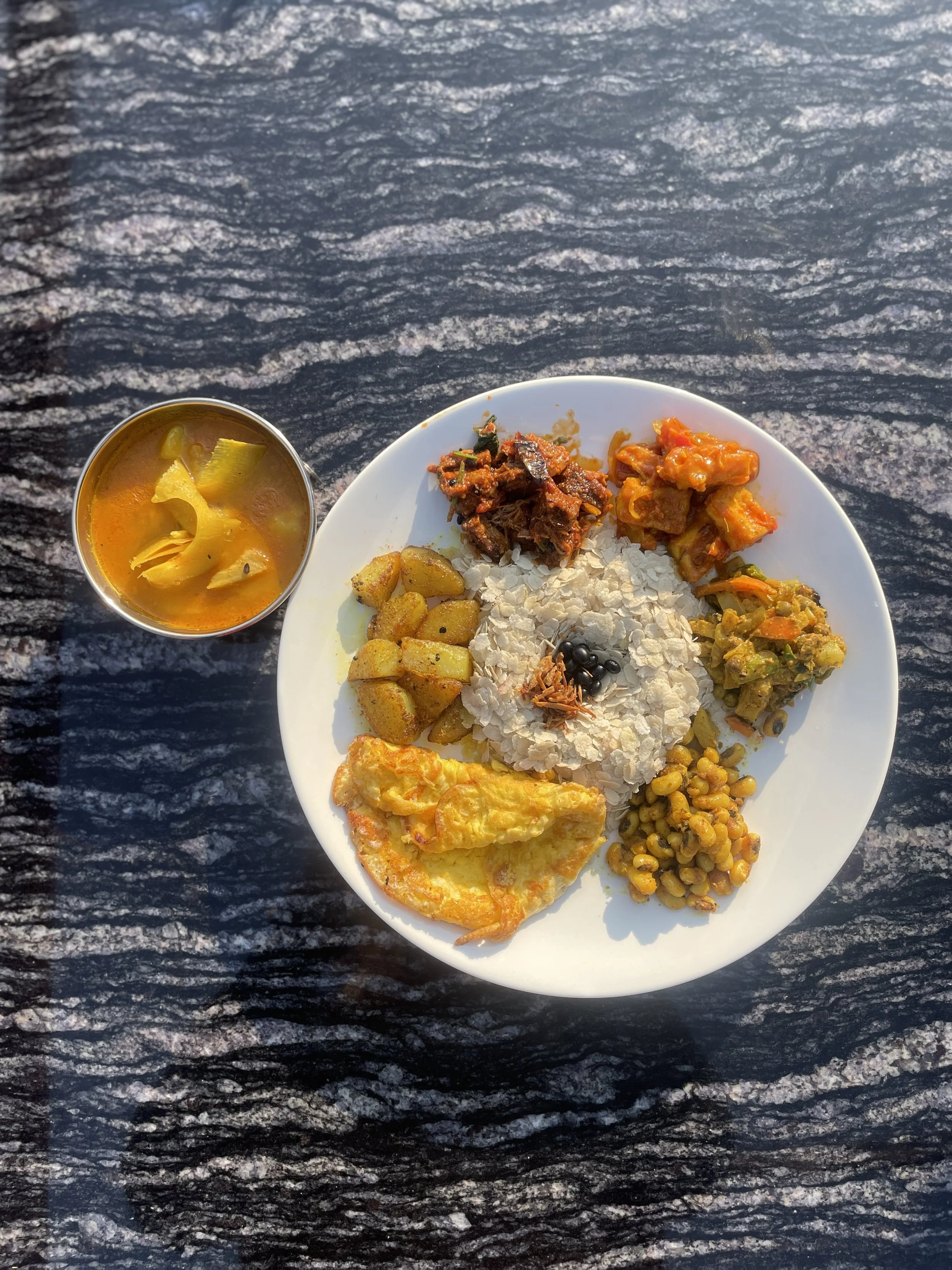 A traditional Newari festival meal with a variety of vegetable and meat dishes served on a white plate with a side of beaten rice and a small bowl of potato and sour bamboo shoot curry, on a black stone table.