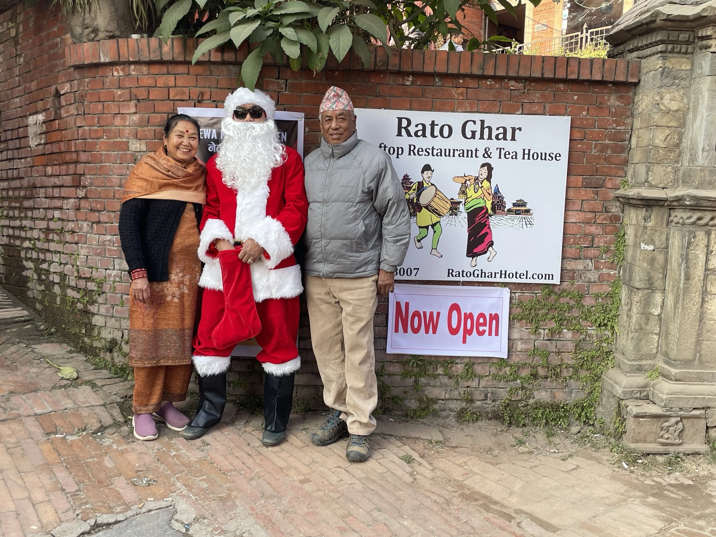 Three people standing outside a restaurant called Rato Ghar, with a sign that says 'Now Open.' The person in the middle is dressed as Santa Claus with sunglasses and a red suit. The woman on the left wears traditional attire, and the man on the right