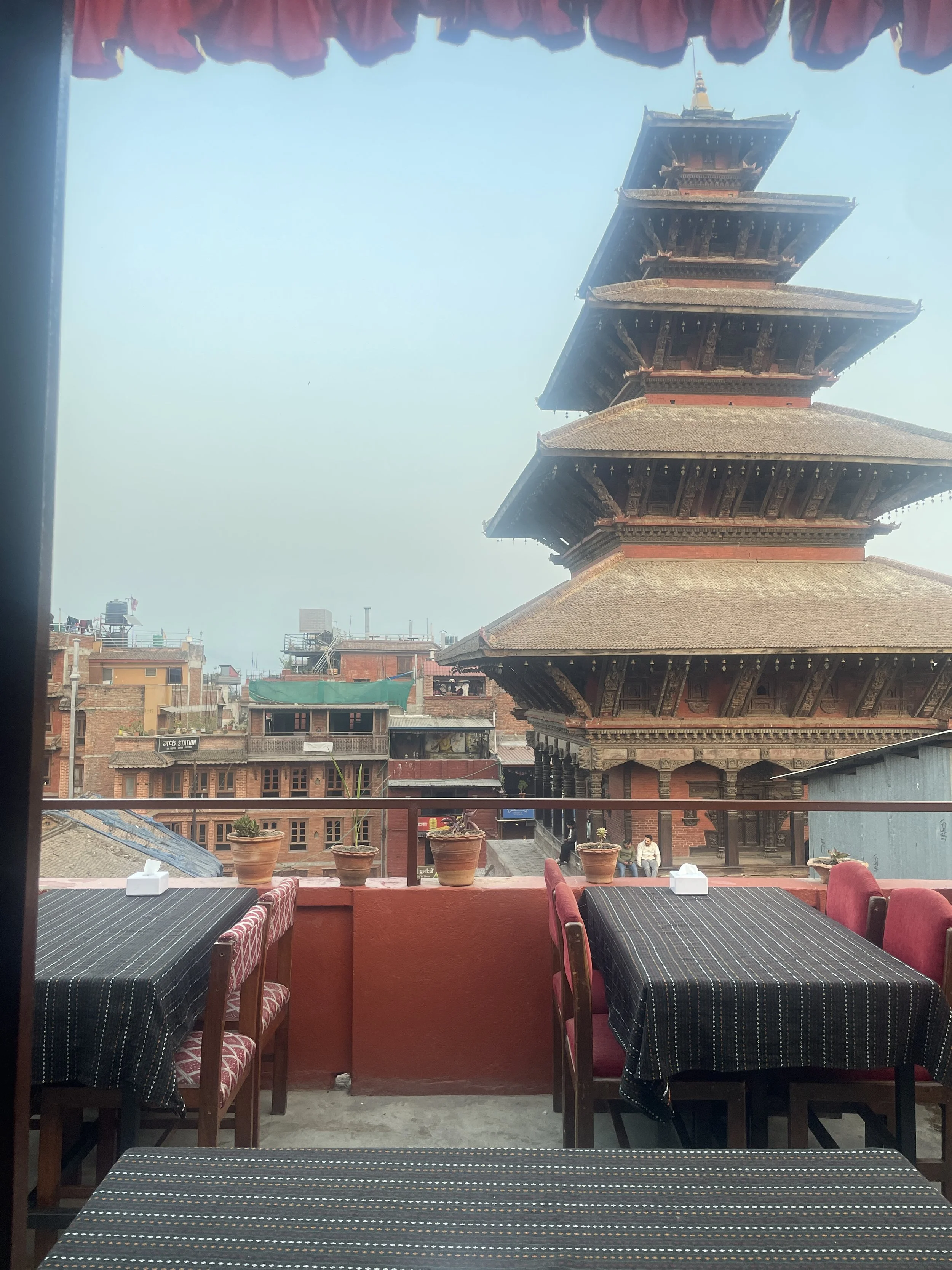 Outdoor rooftop dining area at Rato Ghar Boutique Hotel with tables, red walls, and a clear view of Nyatapola Temple.