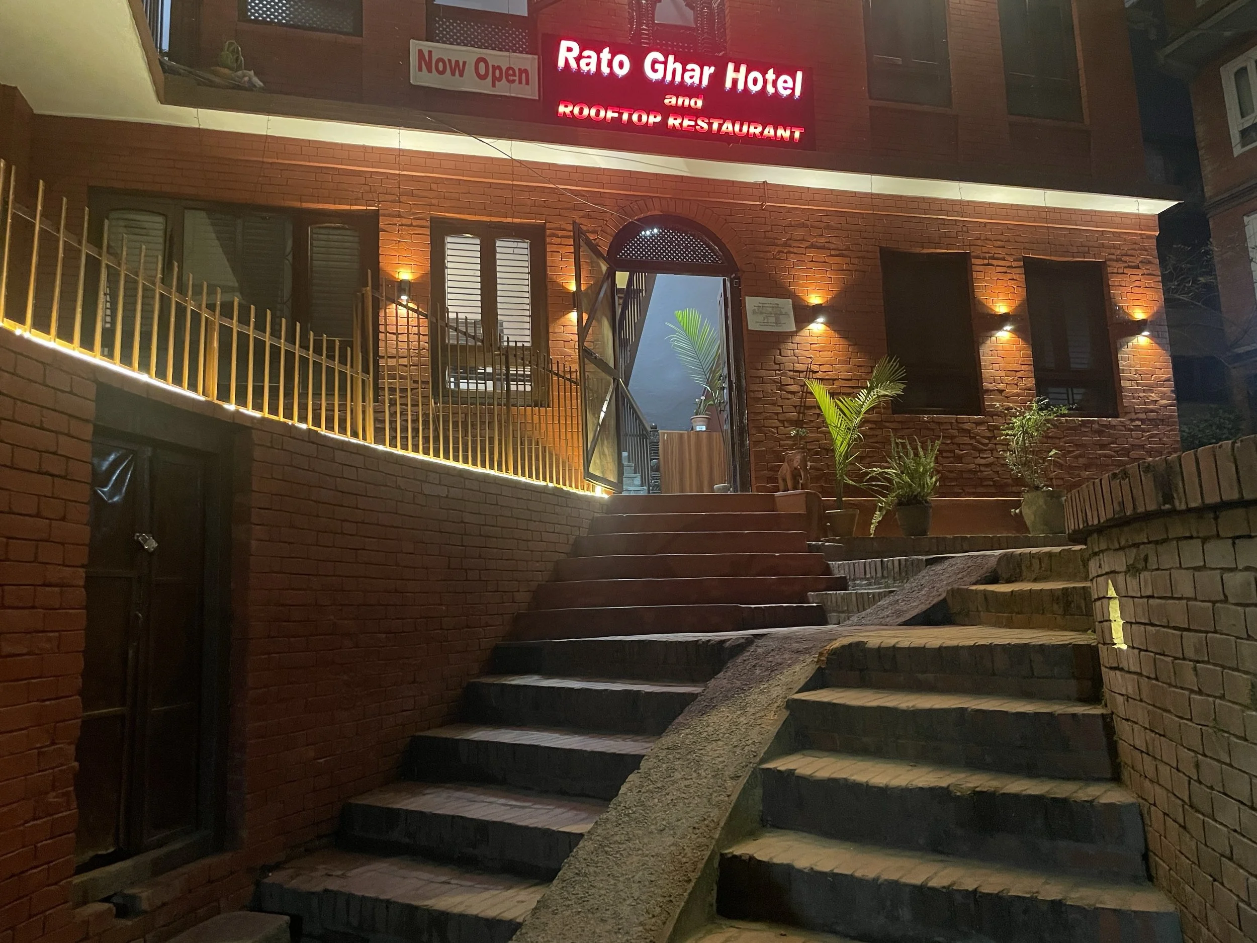 Night view of the entrance to Rato Ghar Hotel and Rooftop Restaurant with a staircase, potted plants, brick walls, and illuminated signs.
