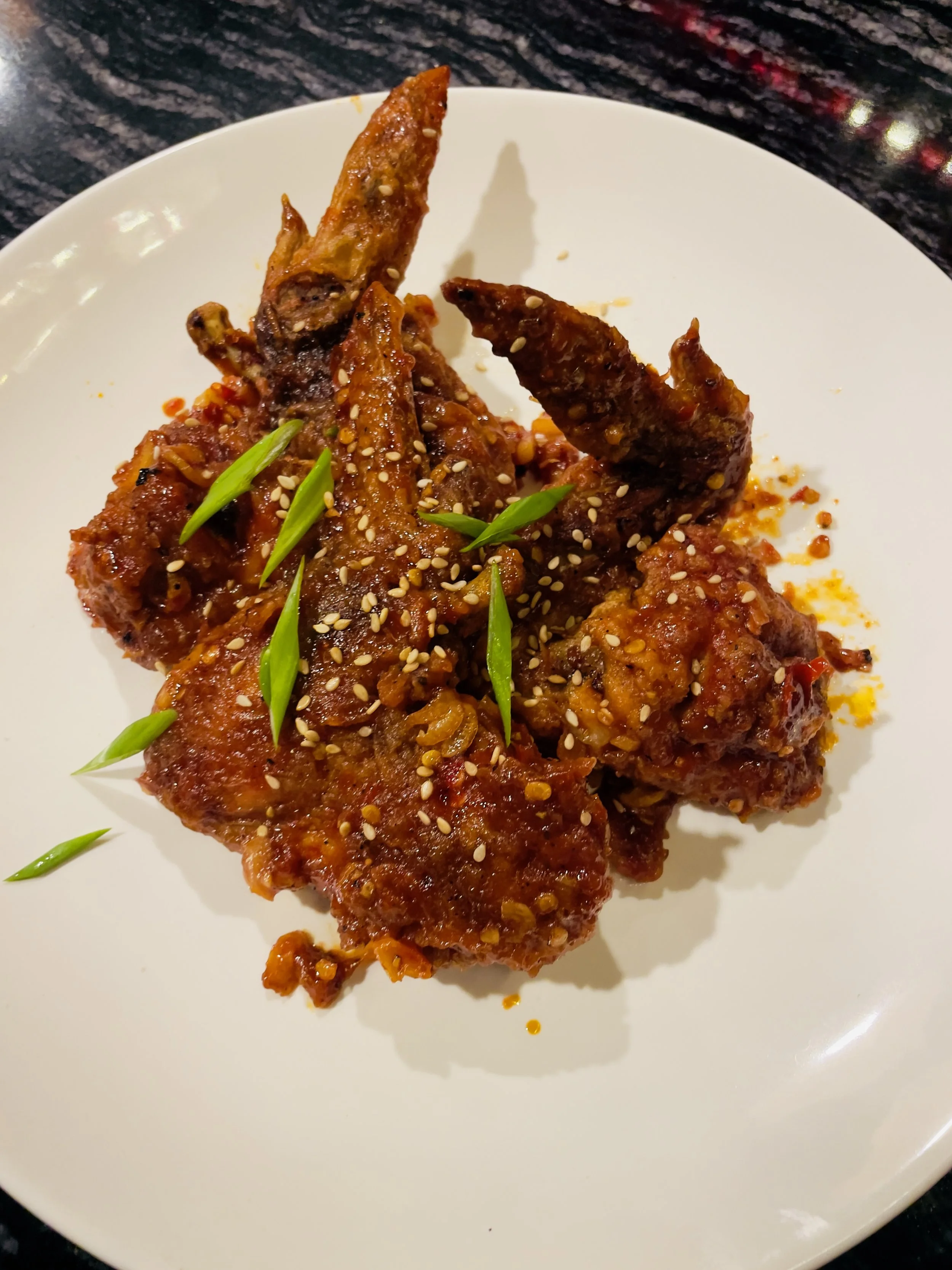 Close-up of spicy glazed chicken wings garnished with sesame seeds and chopped green onions on a white plate.