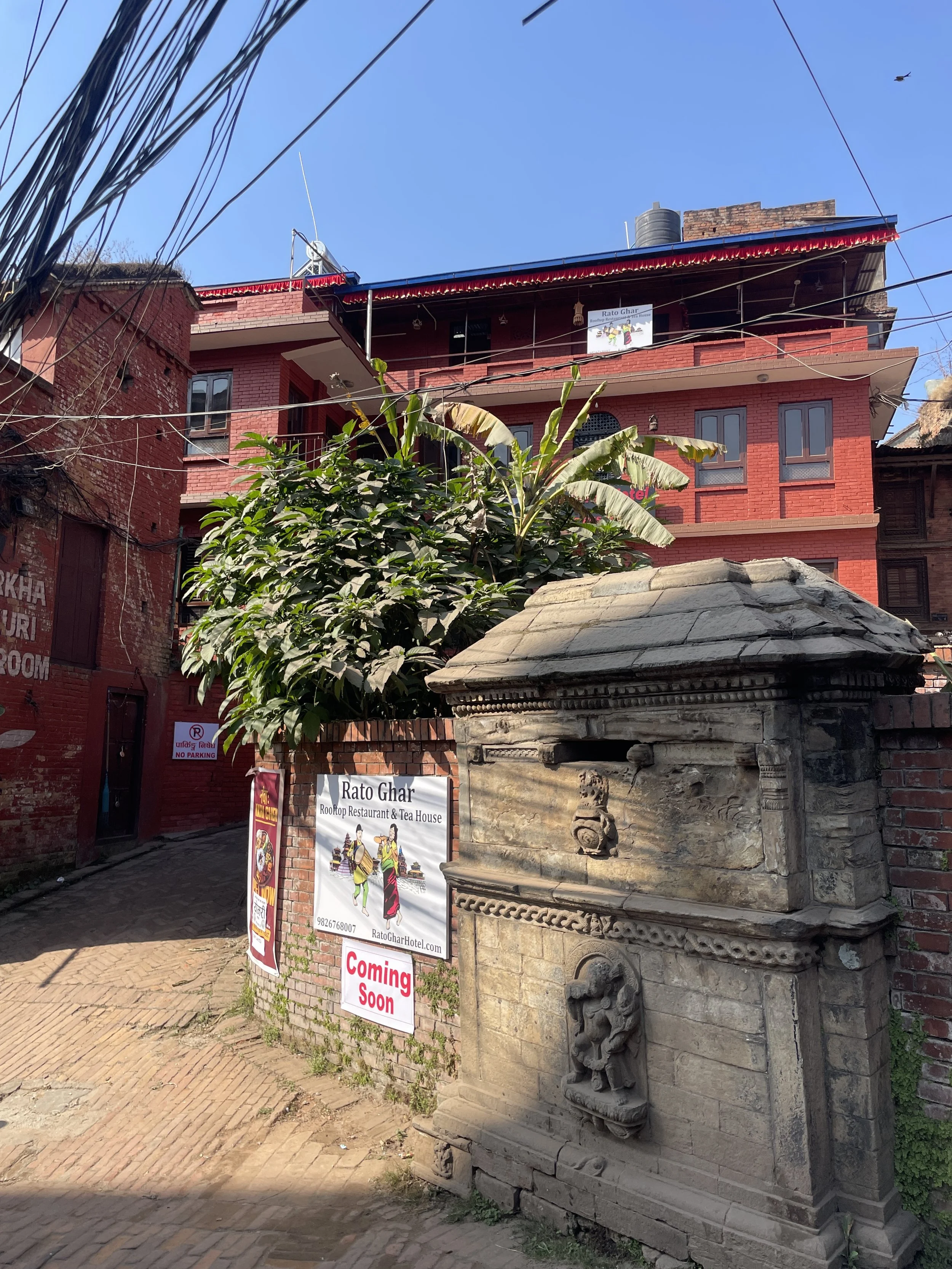 A street scene with a stone shrine in the foreground, a large leafy tree, and red brick buildings in the background. A sign reads 'Rato Ghar Rooftop Restaurant & Tea House' with an illustration of traditional dancers.