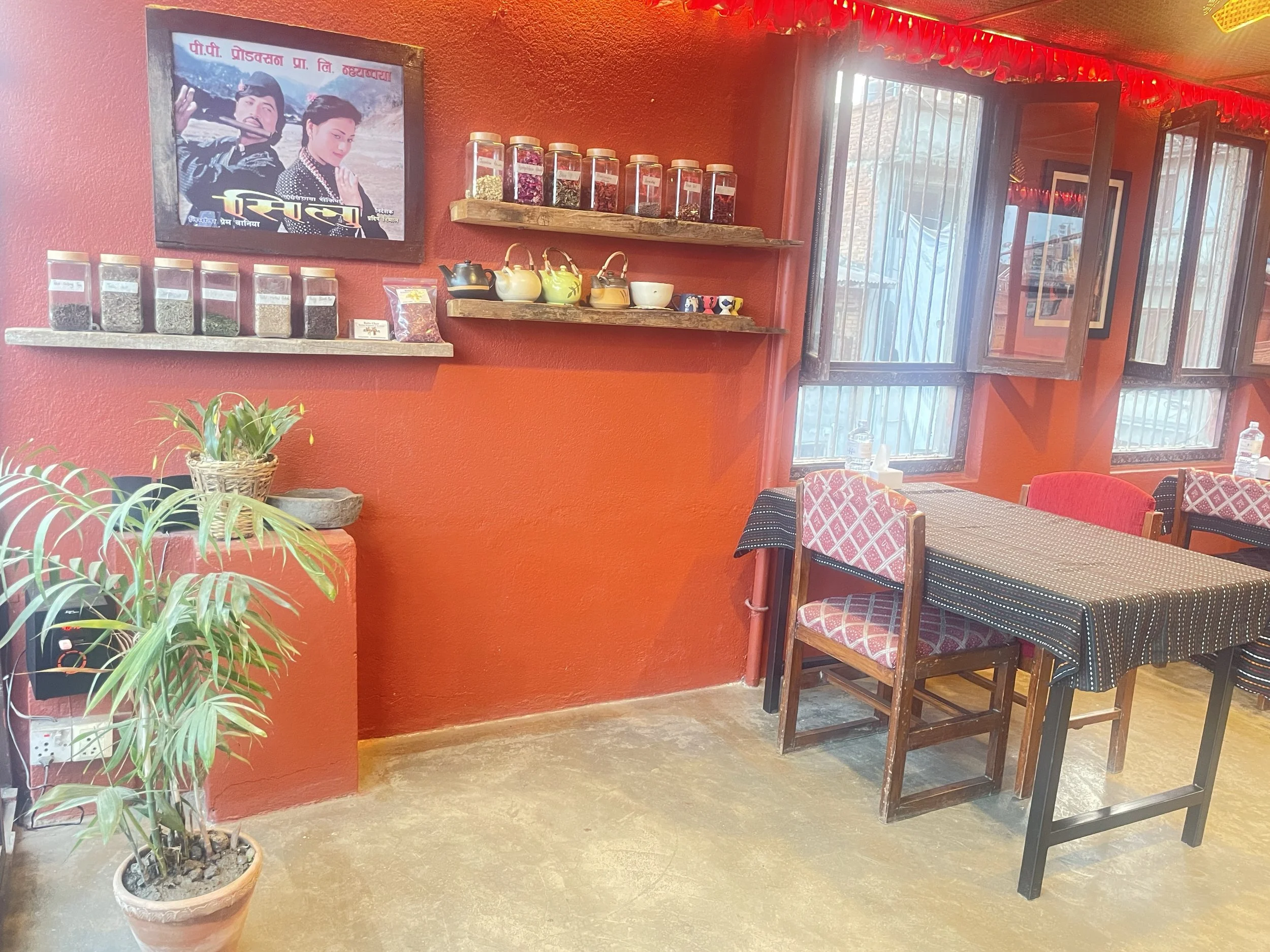 Cozy dining space at Rato Ghar Rooftop Restaurant in Bhaktapur with bright red walls, jars of loose-leaf tea on wooden shelves, teapots, framed artwork, and wooden tables near open windows.