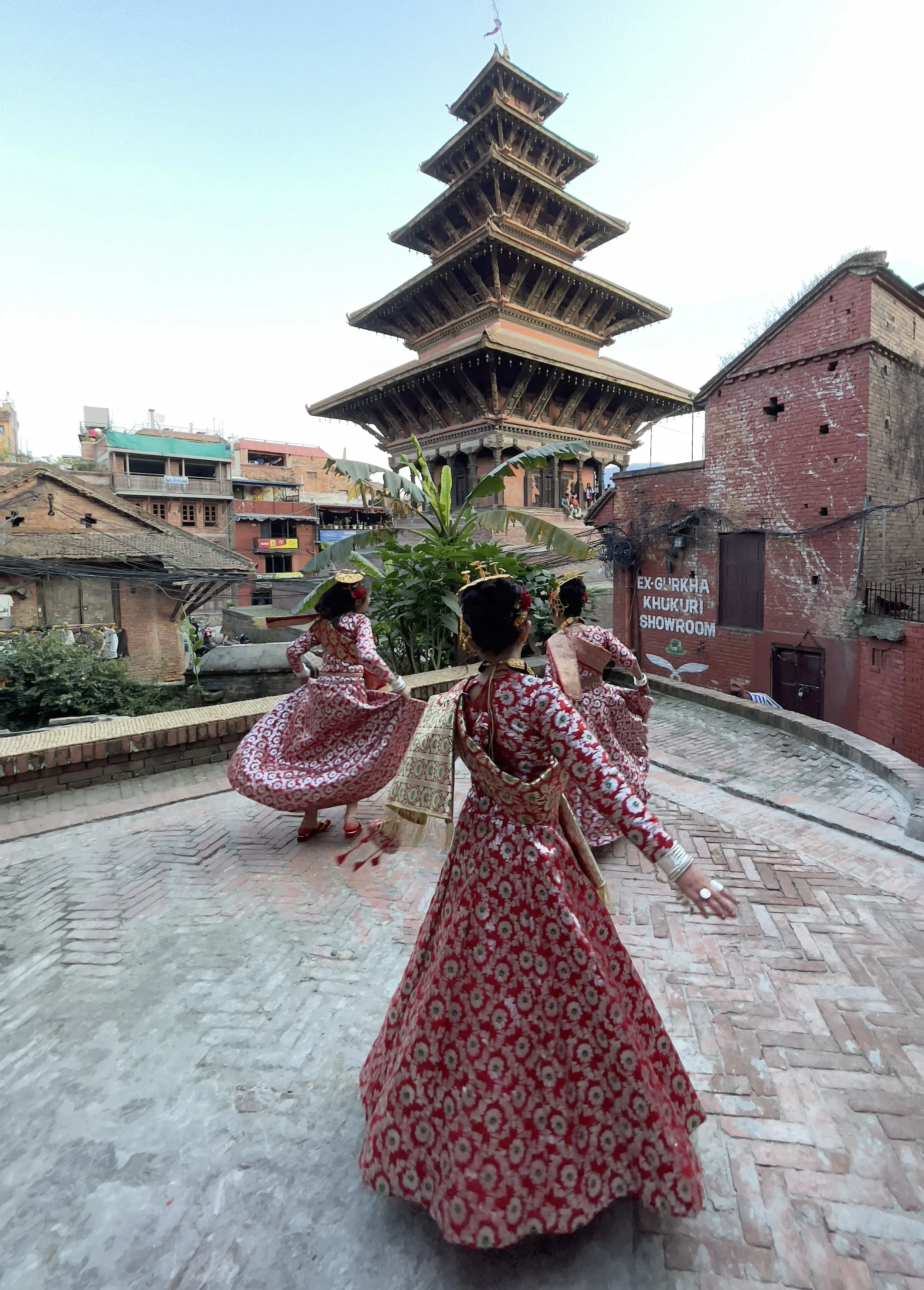 Three women in traditional red and white dresses performing a dance on a brick pathway in front of a pagoda-style building, with a surrounding village and a sign that reads 'Ex-Gurkha Khukuri Showroom'.