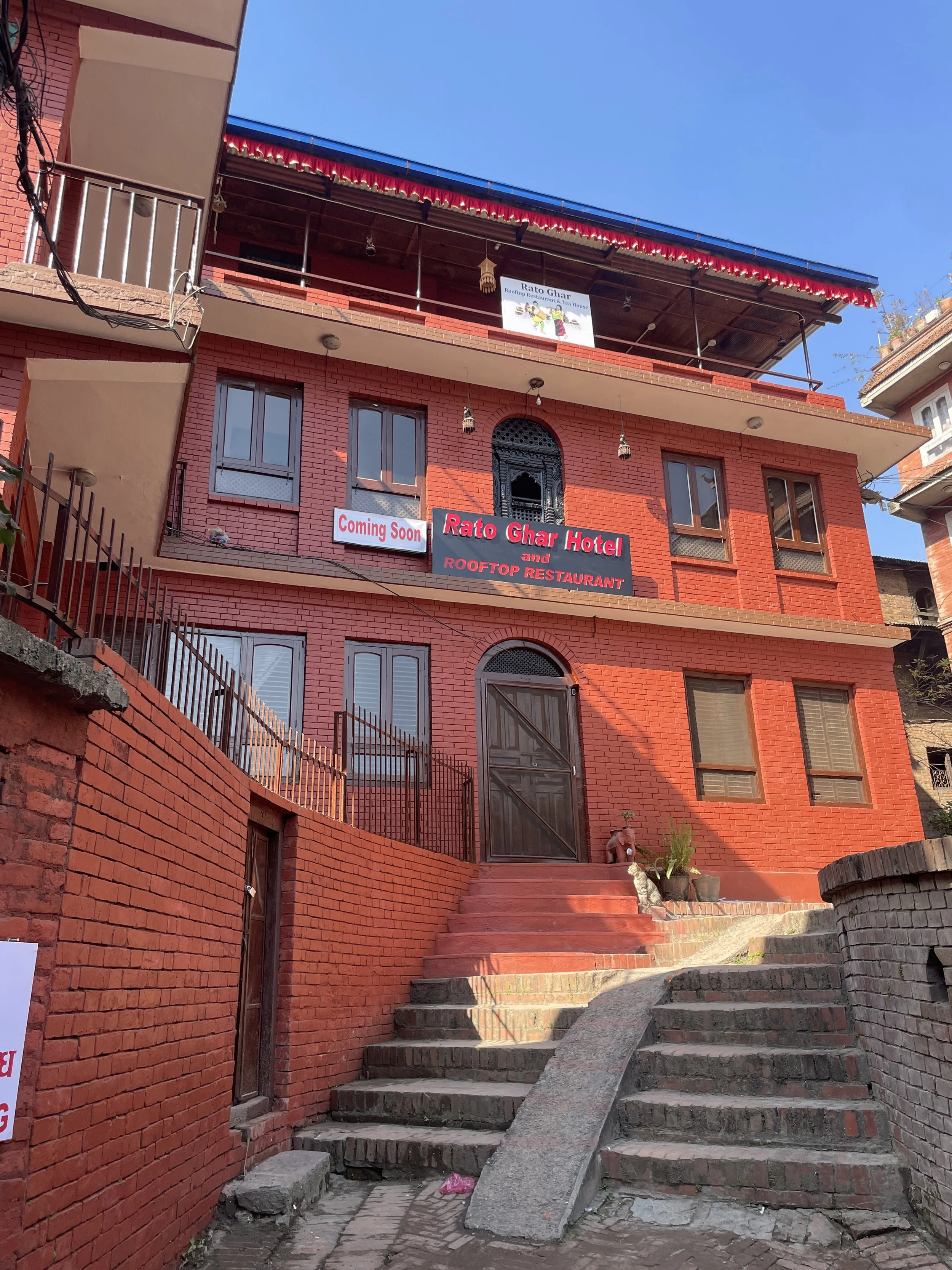 A three-story red brick building with a sign indicating it is a hotel and rooftop restaurant named 'Rato Ghar Hotel.' There is a staircase leading up to the entrance, which is flanked by potted plants. A sign on the second floor reads 'Coming Soon.' 