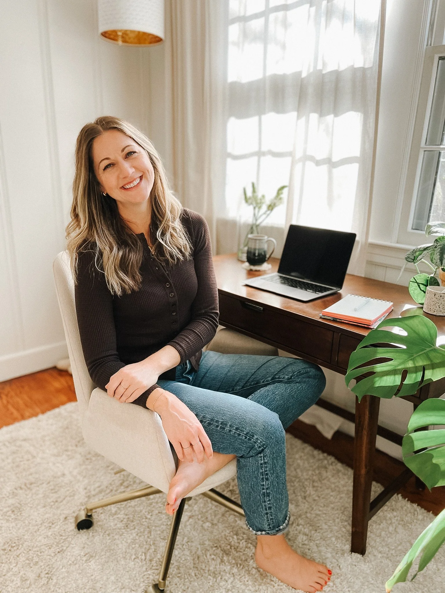 A woman with long, wavy blonde hair sitting in a beige office chair, smiling. She is barefoot, wearing a black long-sleeve top and blue jeans. She is positioned next to a wooden desk with a laptop, notebook, and a coffee pot in a bright, cozy room with large windows and sheer curtains.