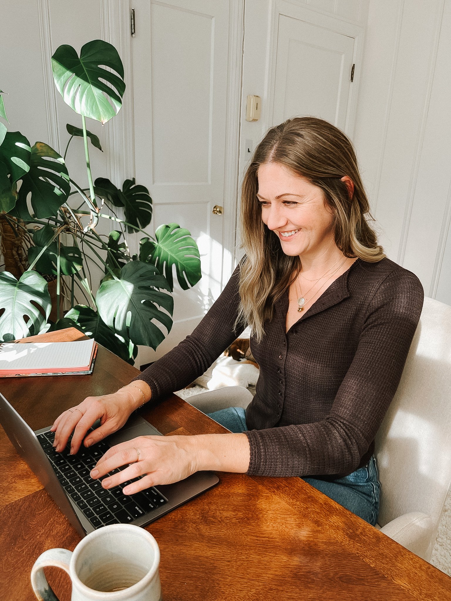 A woman sitting at a wooden table typing on a laptop, with large green houseplants and white doors in the background.