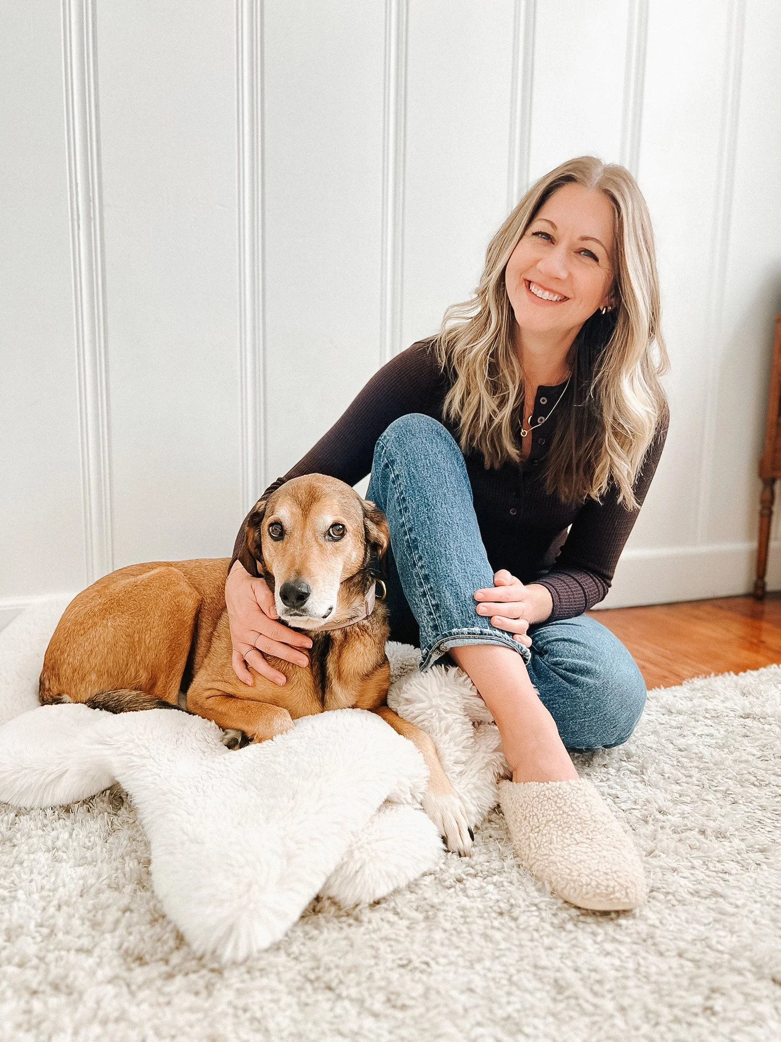 A woman sitting on a plush white rug with a brown dog, smiling and looking at the camera. She is wearing a dark top, blue jeans, and cozy slippers, with a white paneled wall in the background.