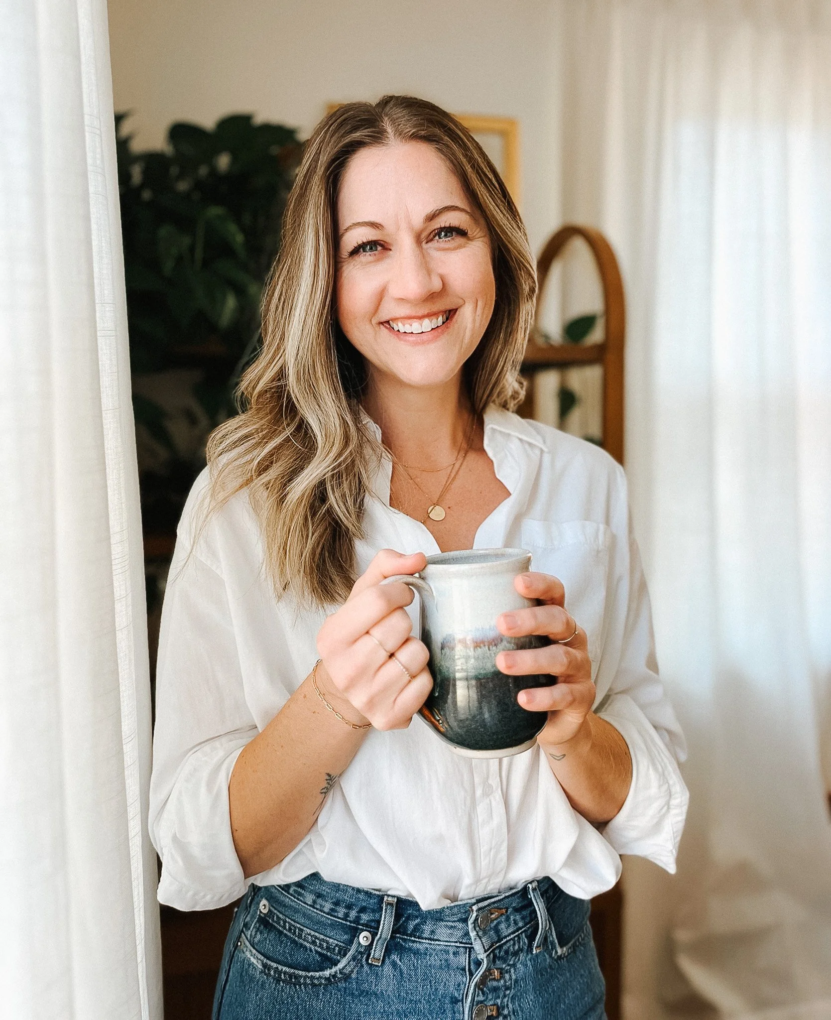 A smiling woman with wavy light brown hair holding a coffee mug, standing in a bright, cozy room.