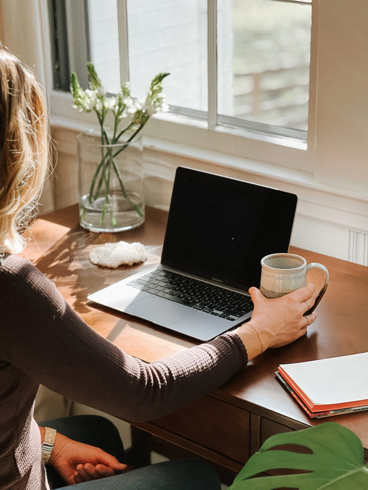 A woman sitting at a wooden desk, holding a mug, with a laptop, notebook, and plant in the background.