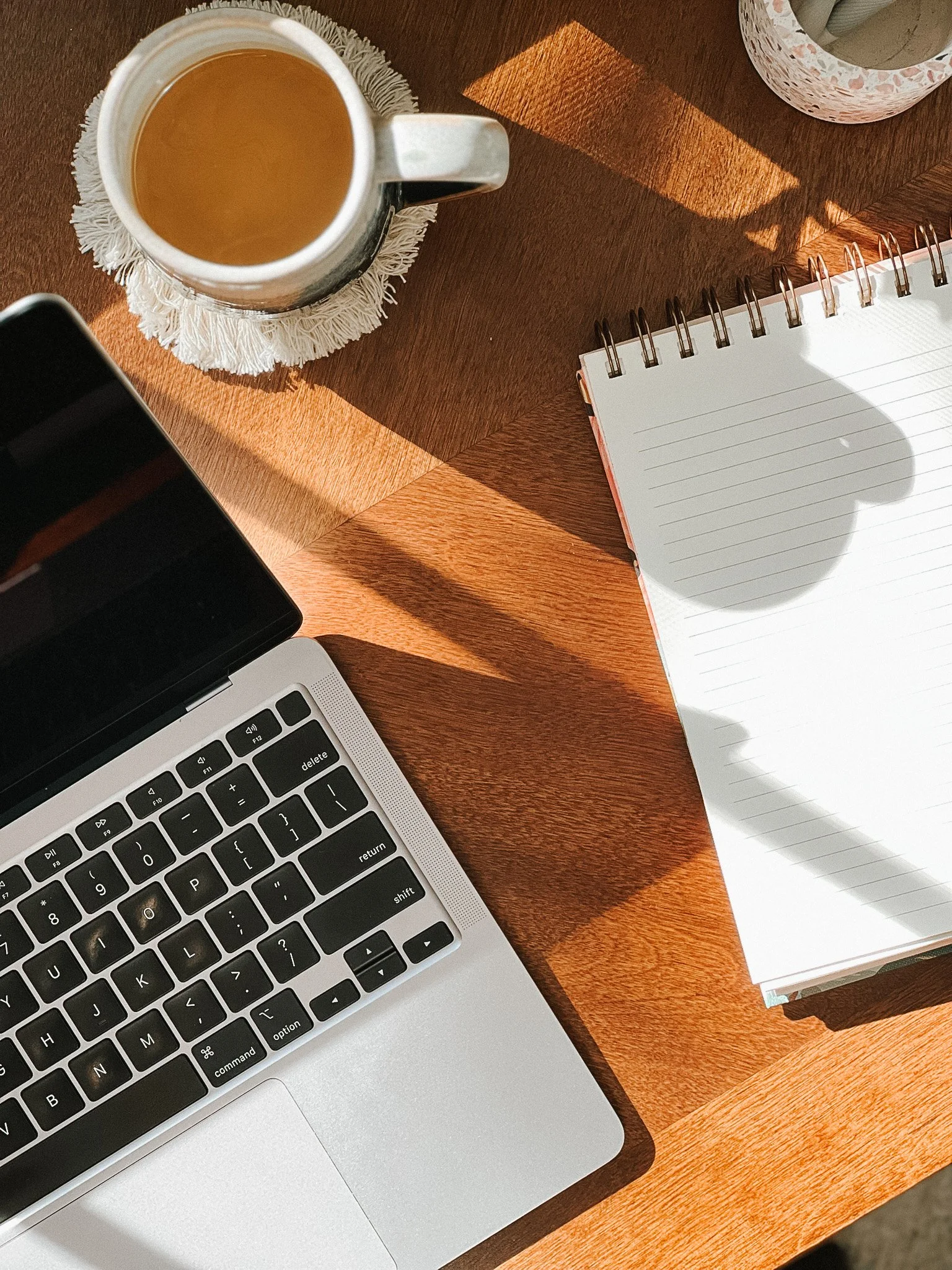 A top view of a wooden desk with a coffee mug on a doily, a silver laptop, and a spiral-bound notebook with lined paper, with sunlight casting shadows.